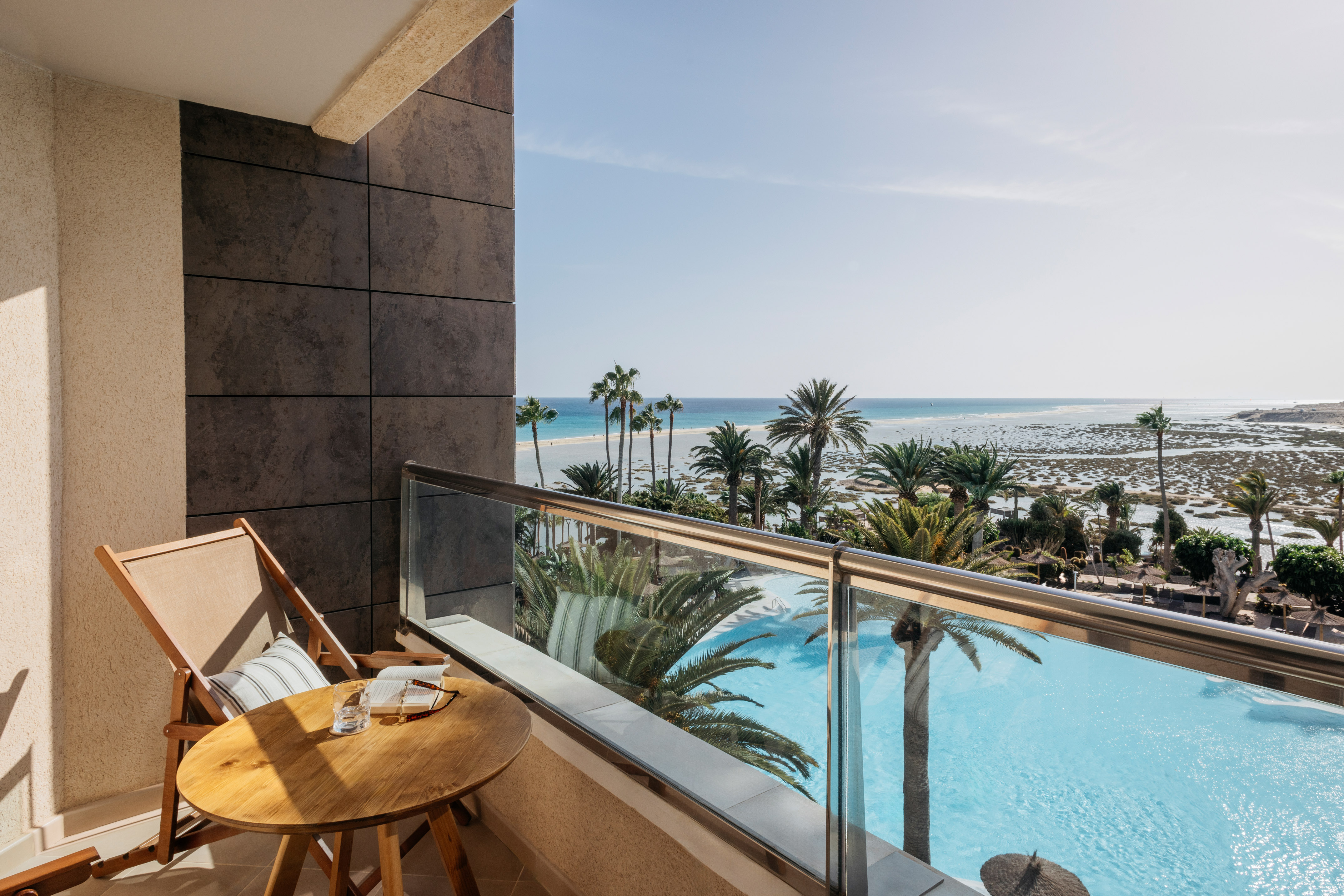 Sunny balcony overlooking tropical ocean, palm trees, and resort pool.