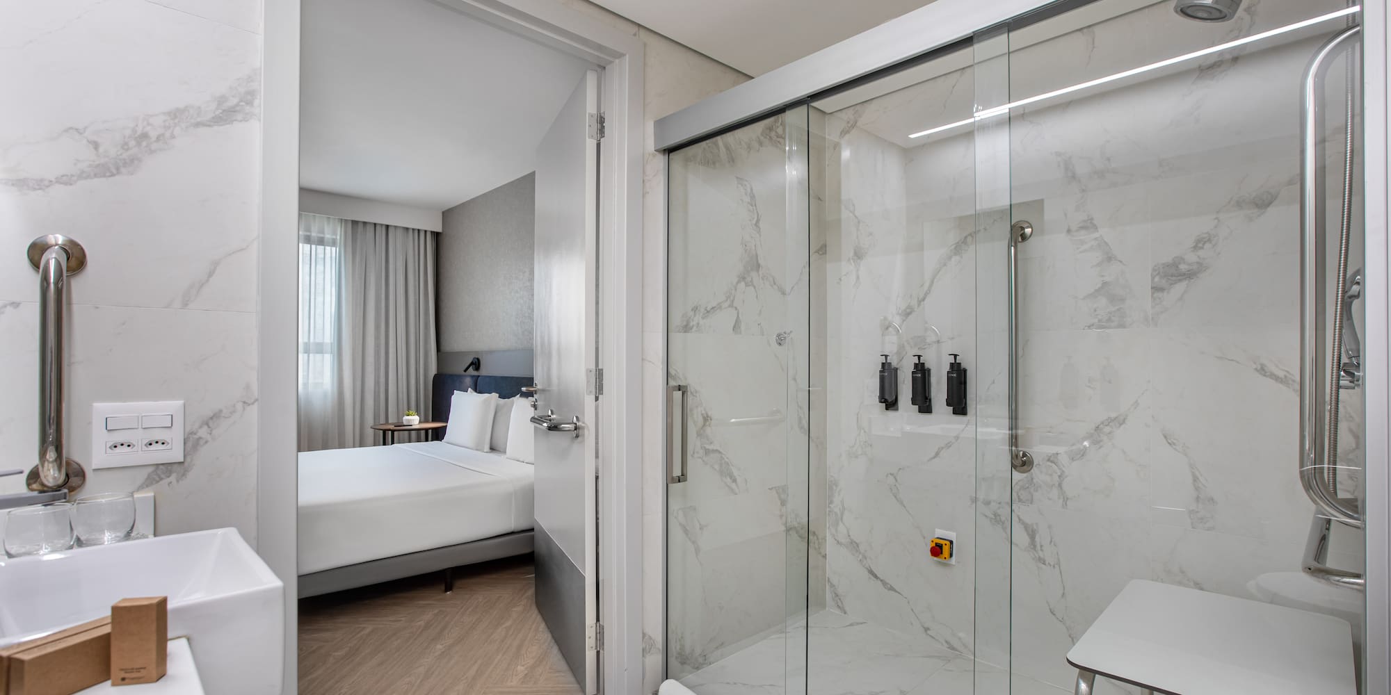 Modern accessible hotel bathroom with marble shower, grab bars, and bedroom view.