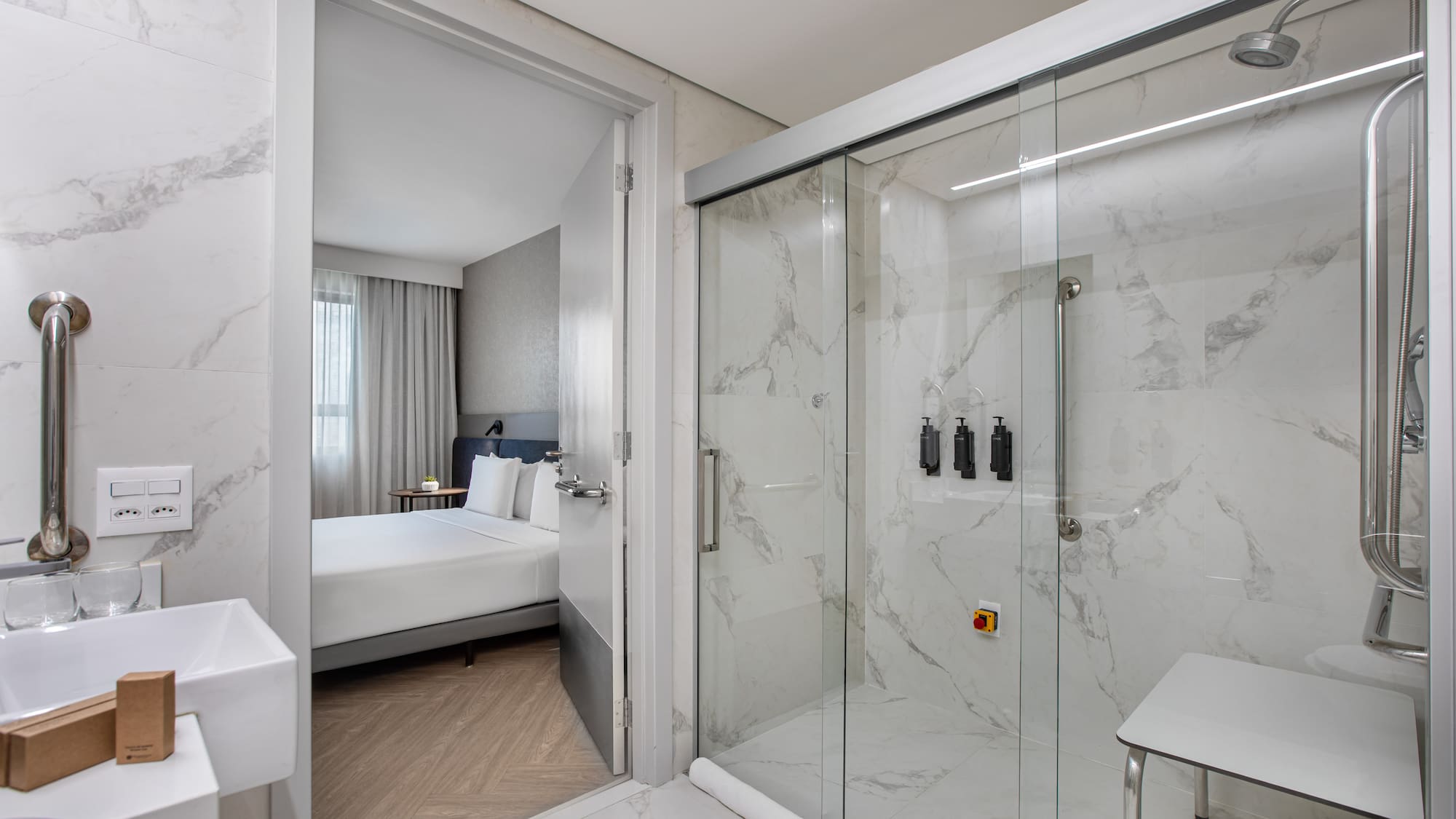 Modern accessible hotel bathroom with marble shower, grab bars, and bedroom view.