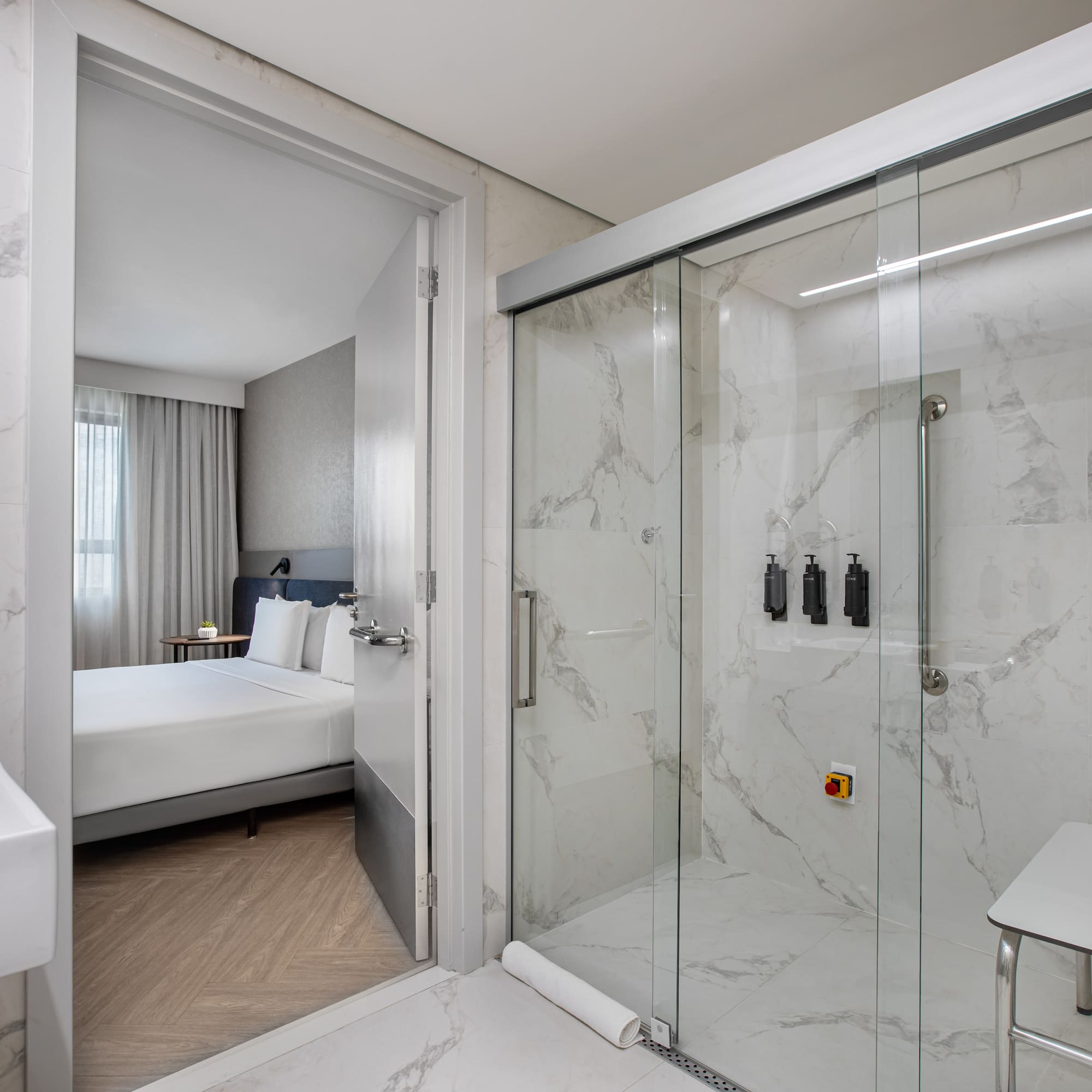 Modern accessible hotel bathroom with marble shower, grab bars, and bedroom view.