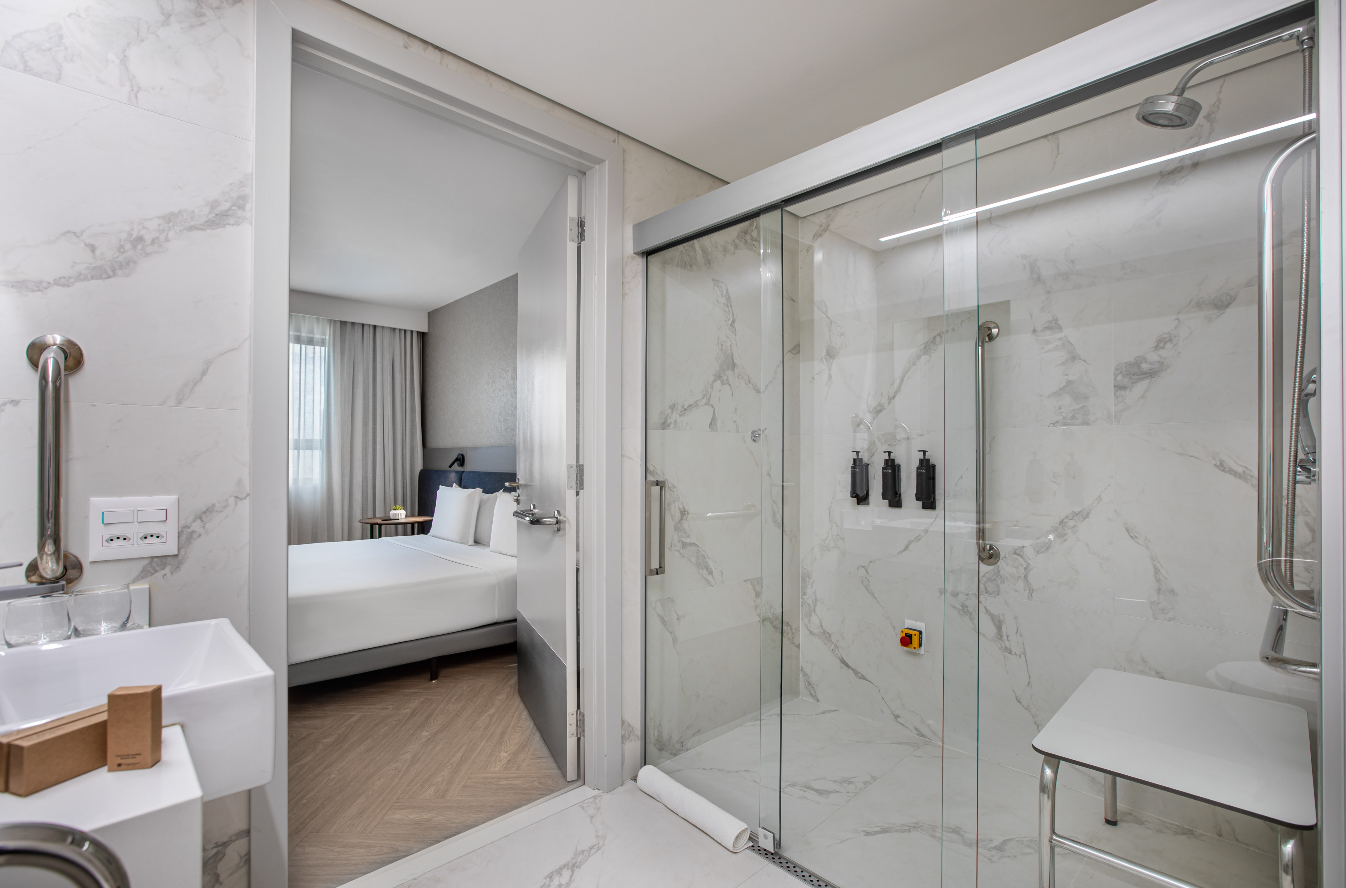 Modern accessible hotel bathroom with marble shower, grab bars, and bedroom view.