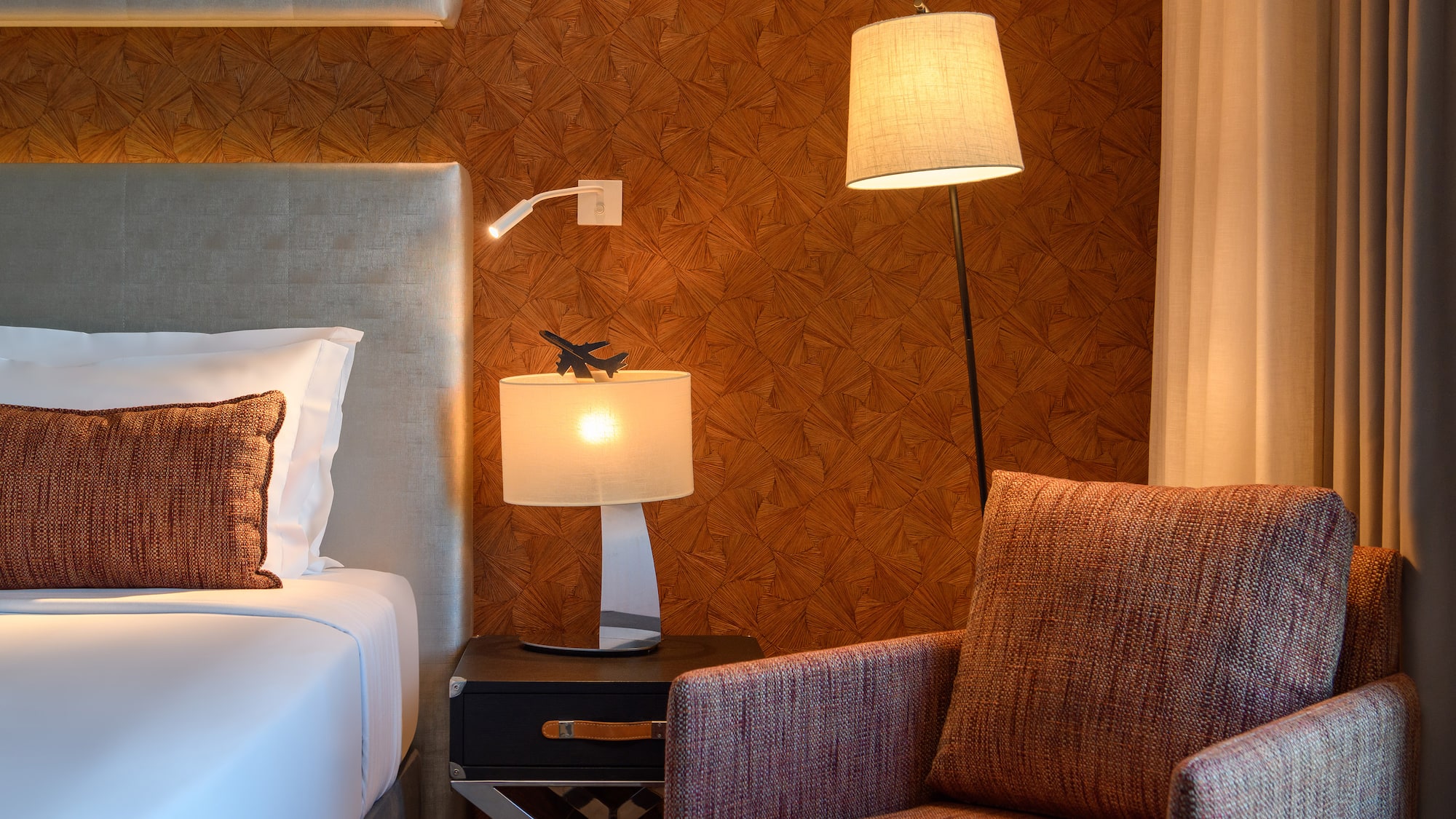 Hotel room with bed, armchair, floor lamp, table lamp, geometric wallpaper.