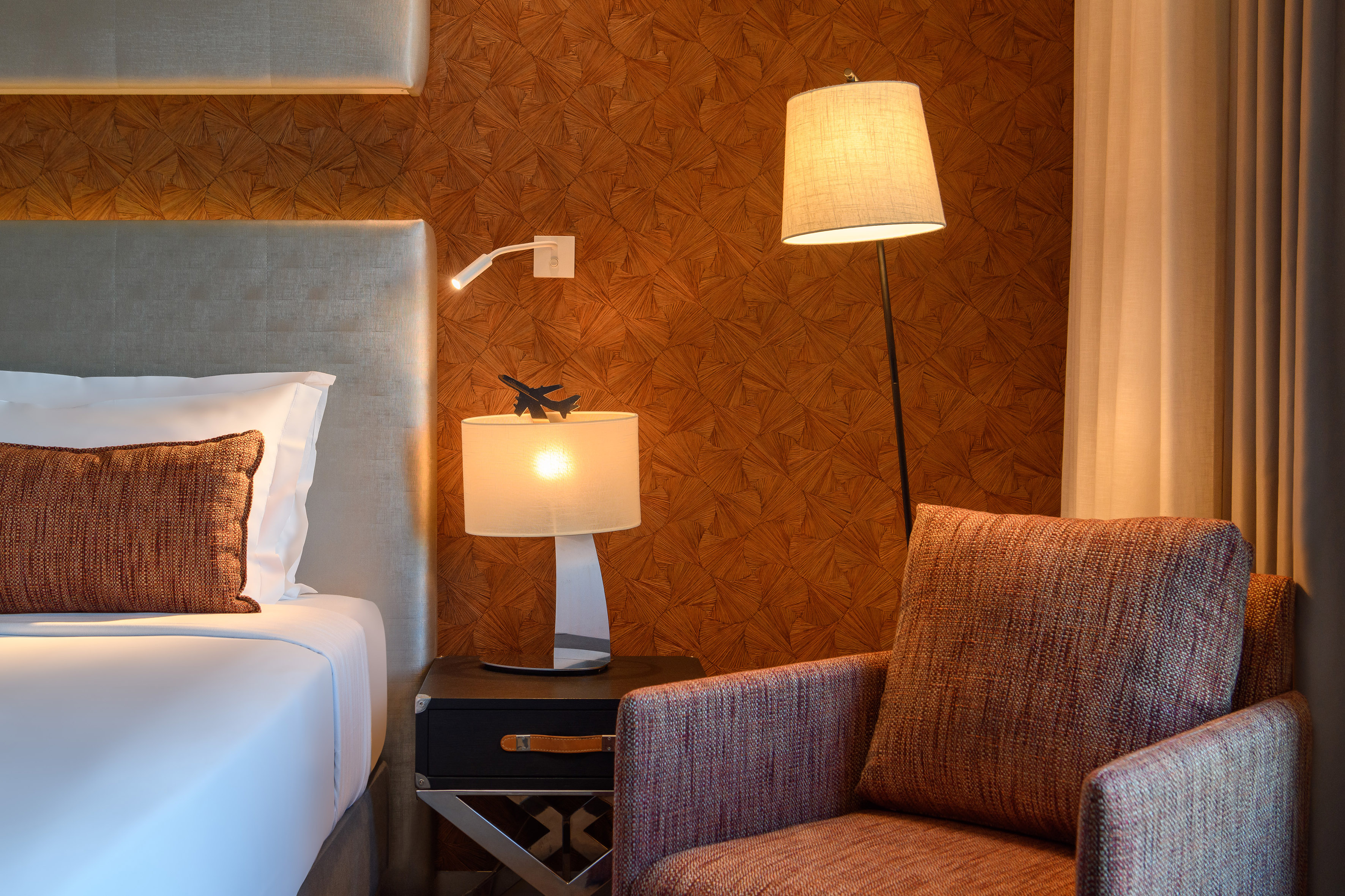 Hotel room with bed, armchair, floor lamp, table lamp, geometric wallpaper.