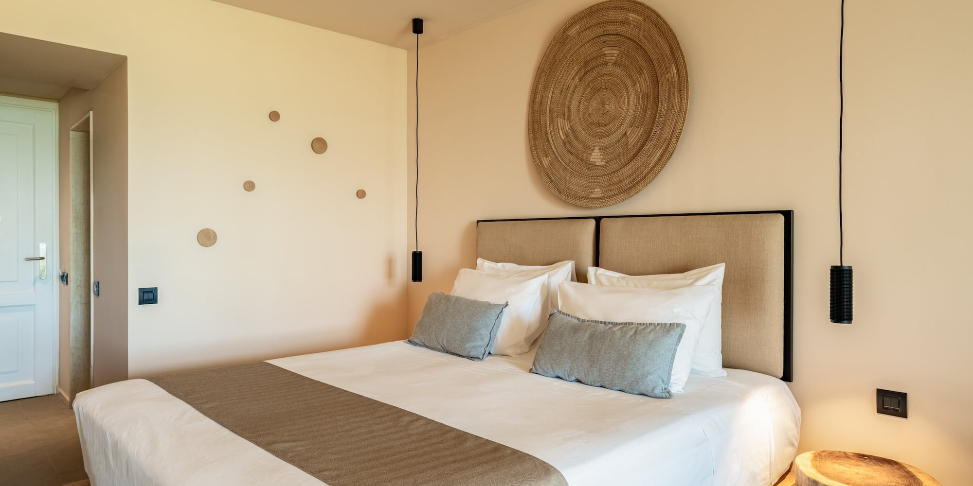 Modern hotel room with king bed, neutral decor, woven art, pendant lights.