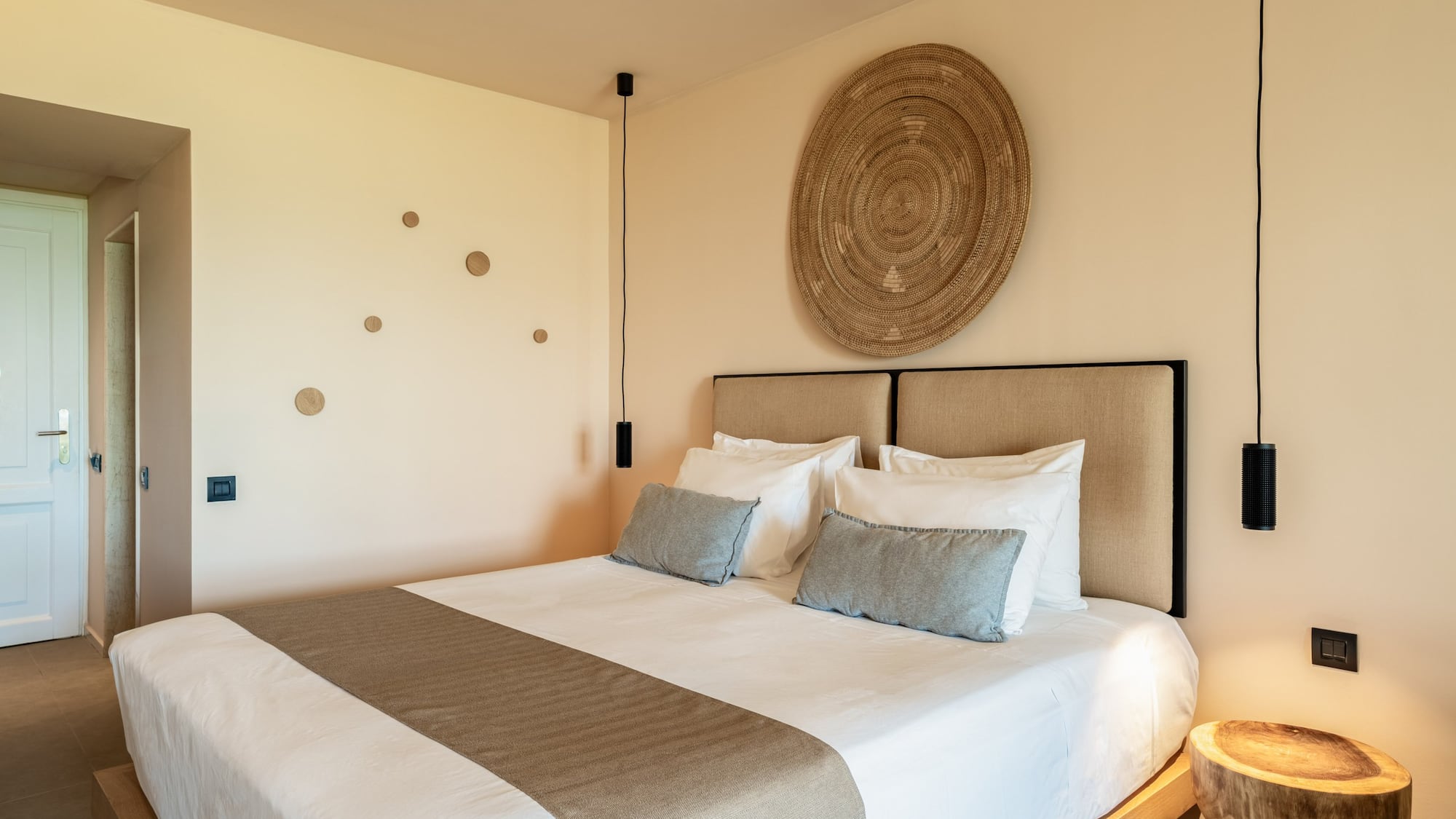 Modern hotel room with king bed, neutral decor, woven art, pendant lights.