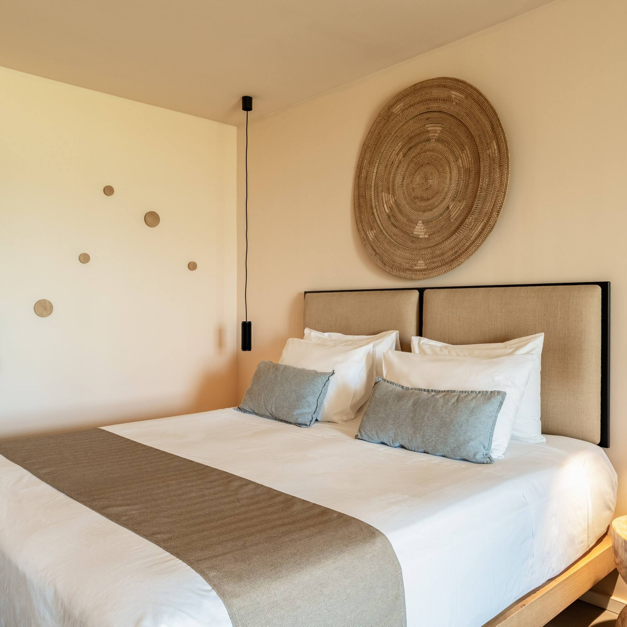 Modern hotel room with king bed, neutral decor, woven art, pendant lights.