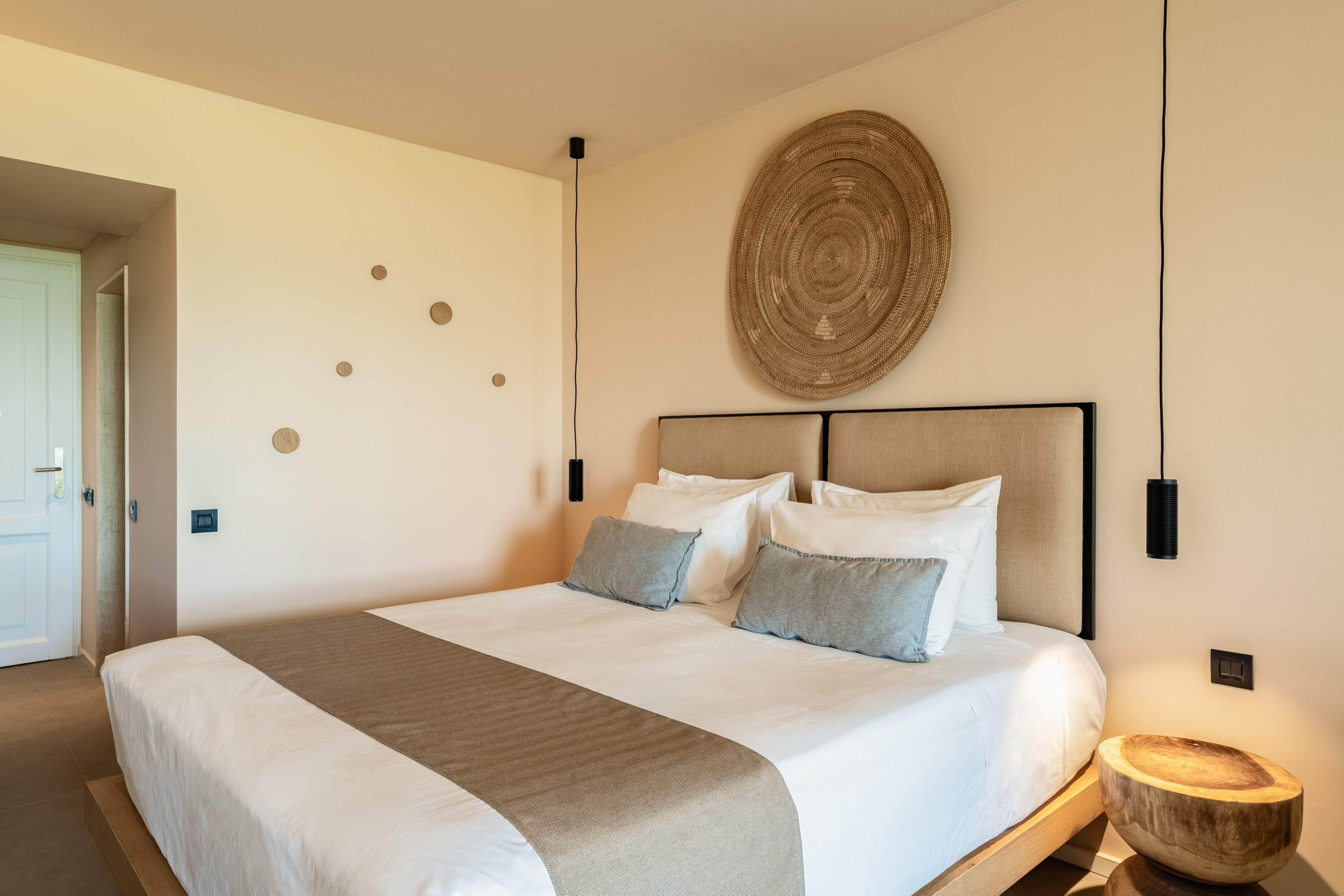 Modern hotel room with king bed, neutral decor, woven art, pendant lights.