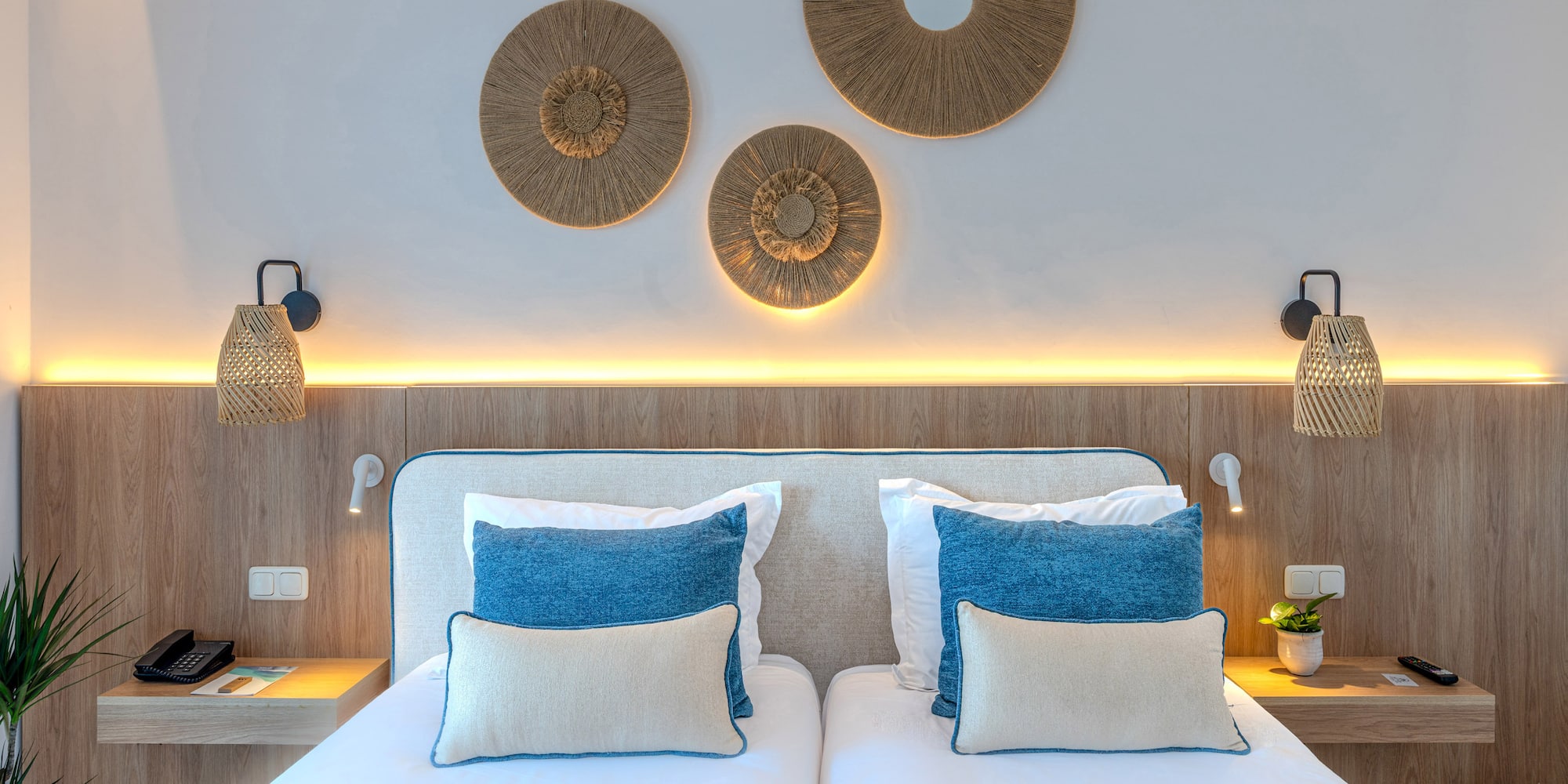 Modern hotel twin bedroom with wooden headboard, blue pillows, and woven decor.