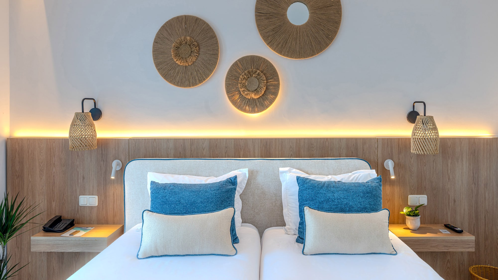 Modern hotel twin bedroom with wooden headboard, blue pillows, and woven decor.