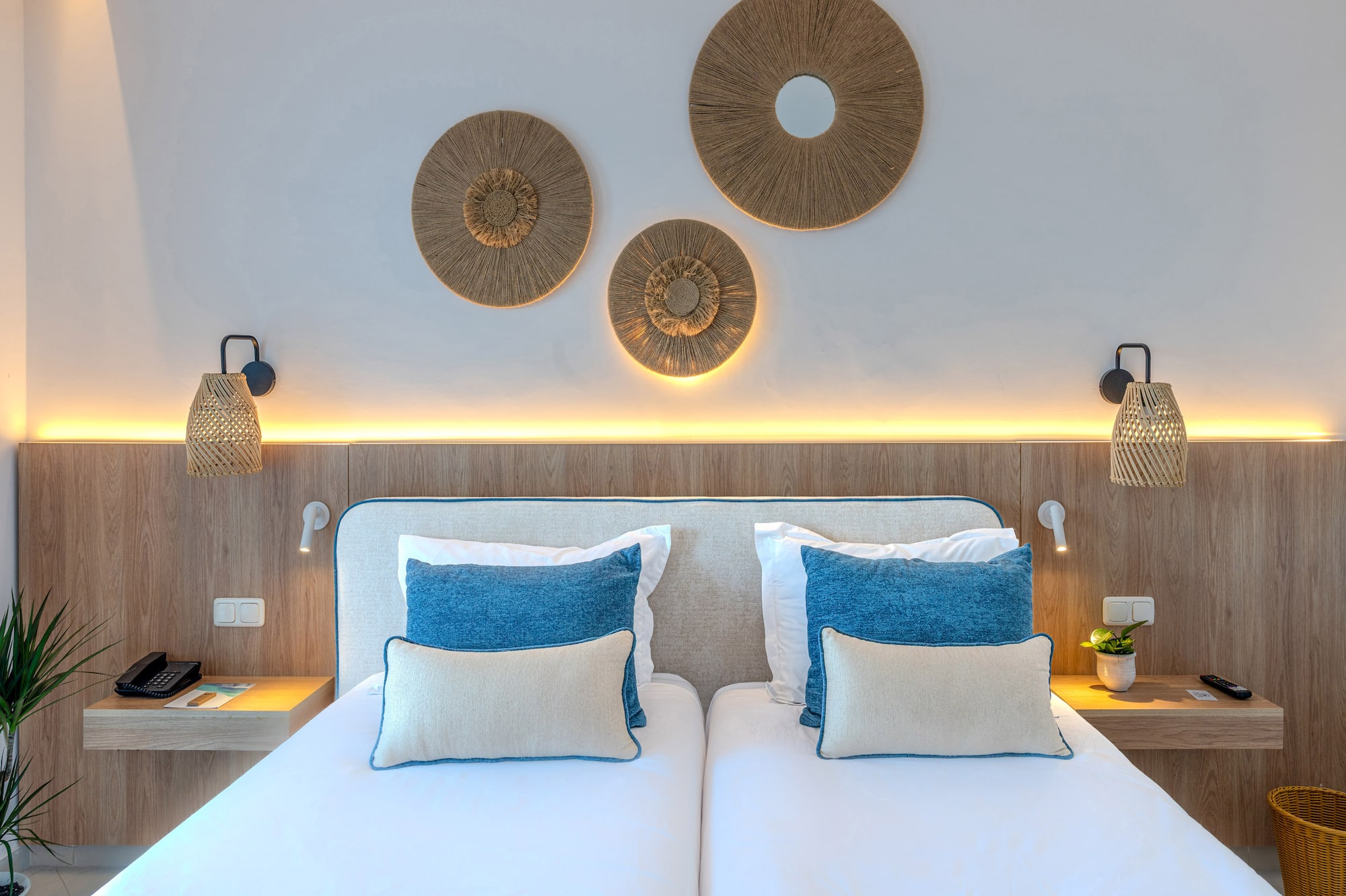 Modern hotel twin bedroom with wooden headboard, blue pillows, and woven decor.