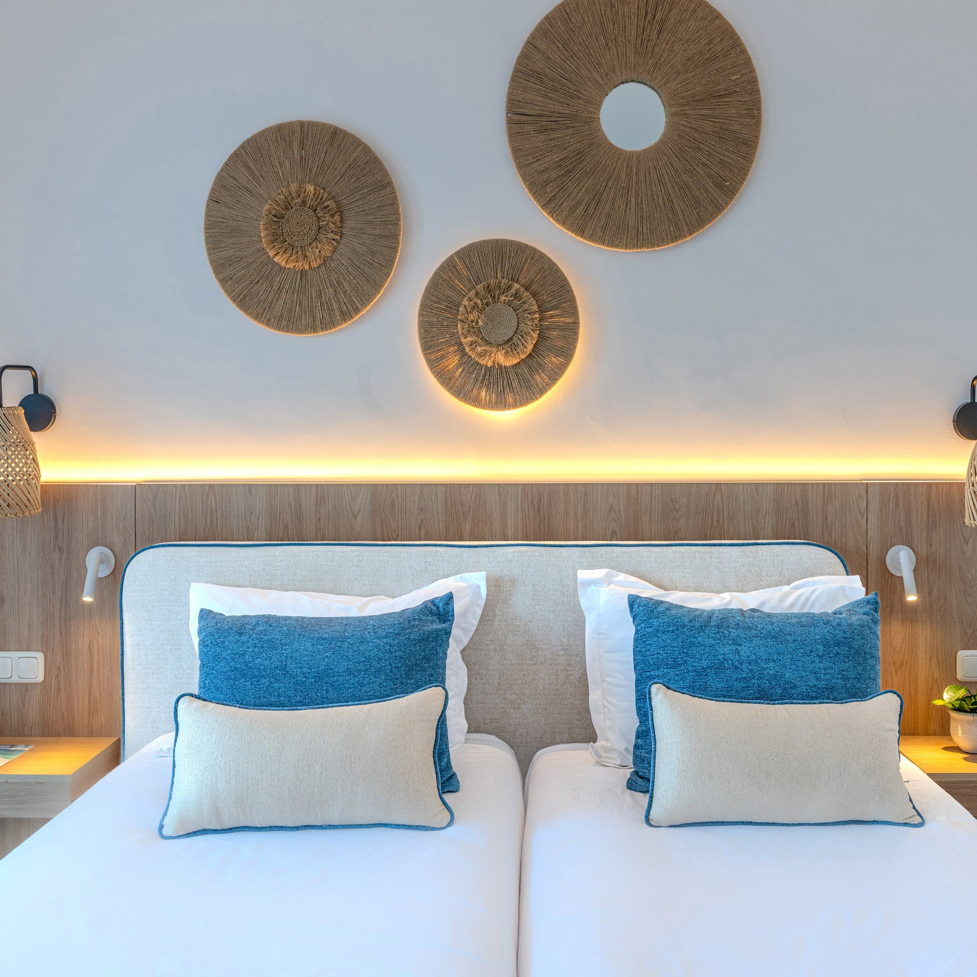 Modern hotel twin bedroom with wooden headboard, blue pillows, and woven decor.