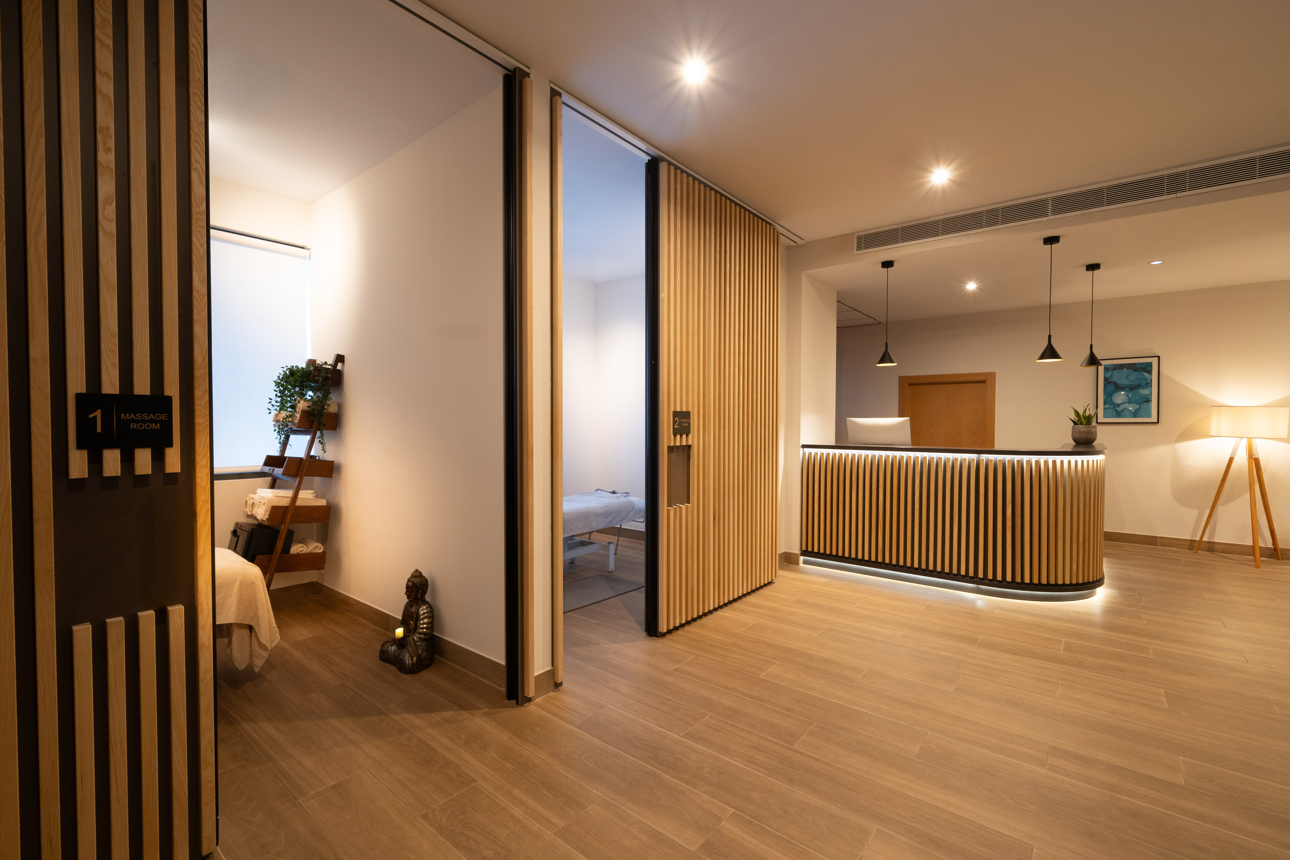 Modern spa reception with wood slats, open massage room, and illuminated desk.