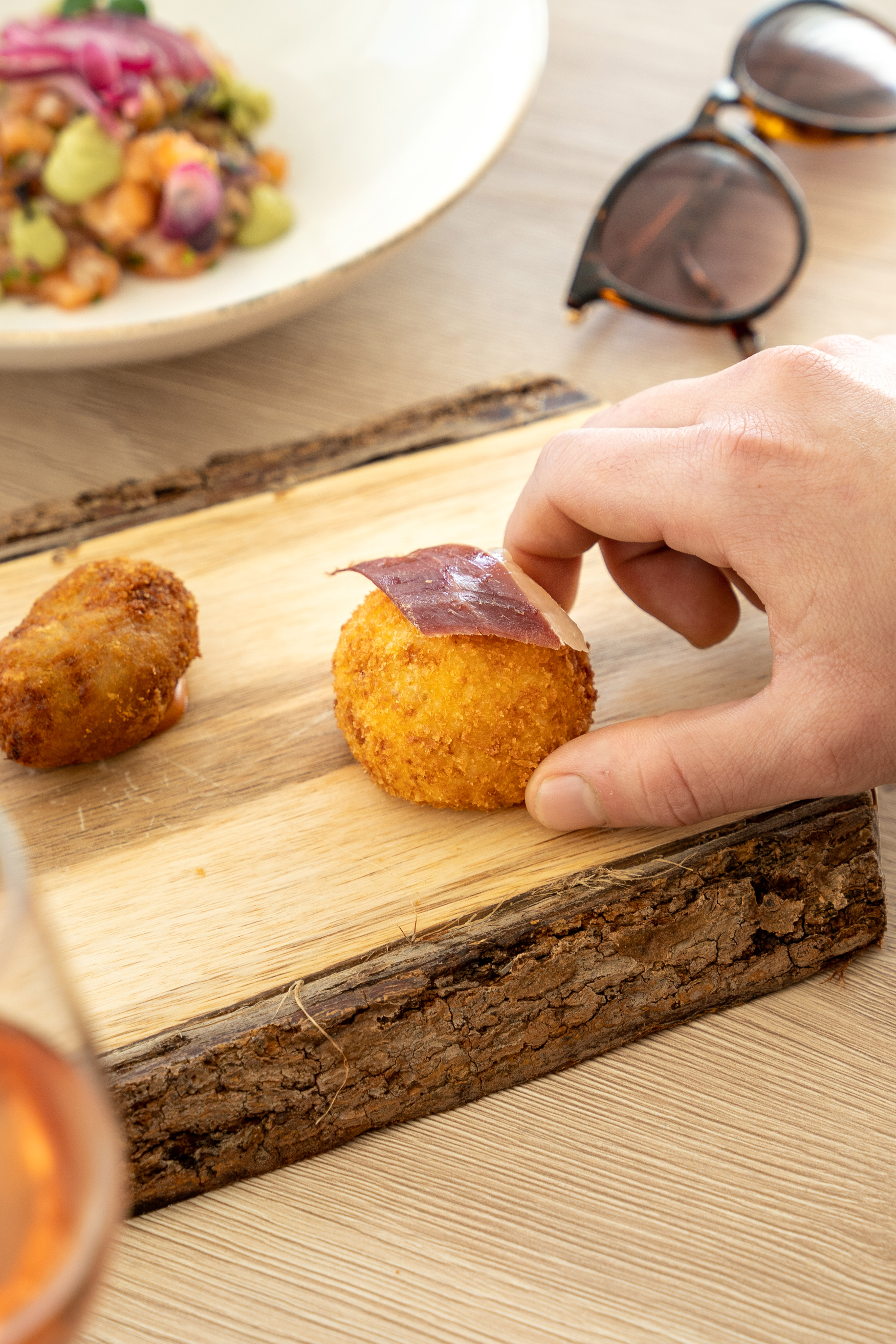 Hand picking a jamón croquette from a rustic wooden board with blurred tapas.
