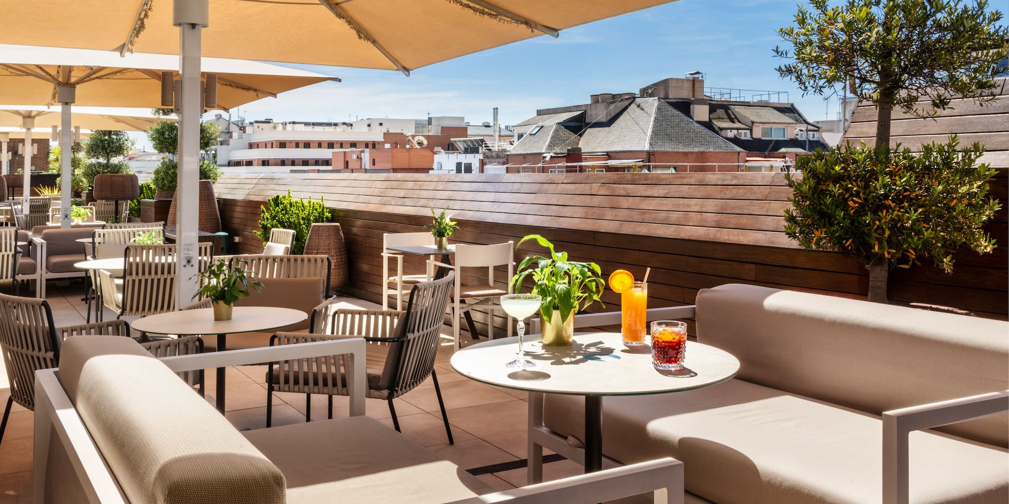 Sunny rooftop terrace with modern outdoor seating, umbrellas, cocktails, and city view.