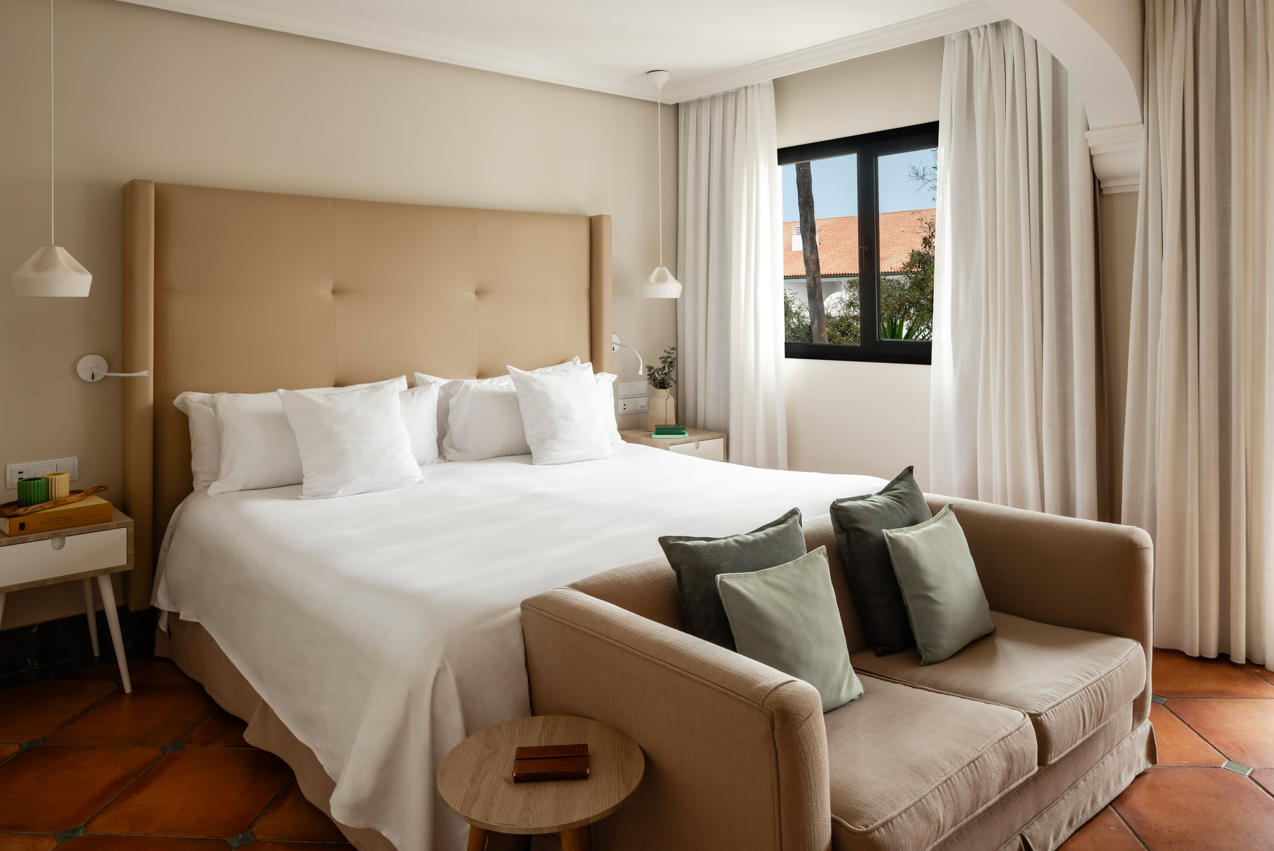 Luxury hotel bedroom with king bed, beige sofa, and terracotta tiled floor.