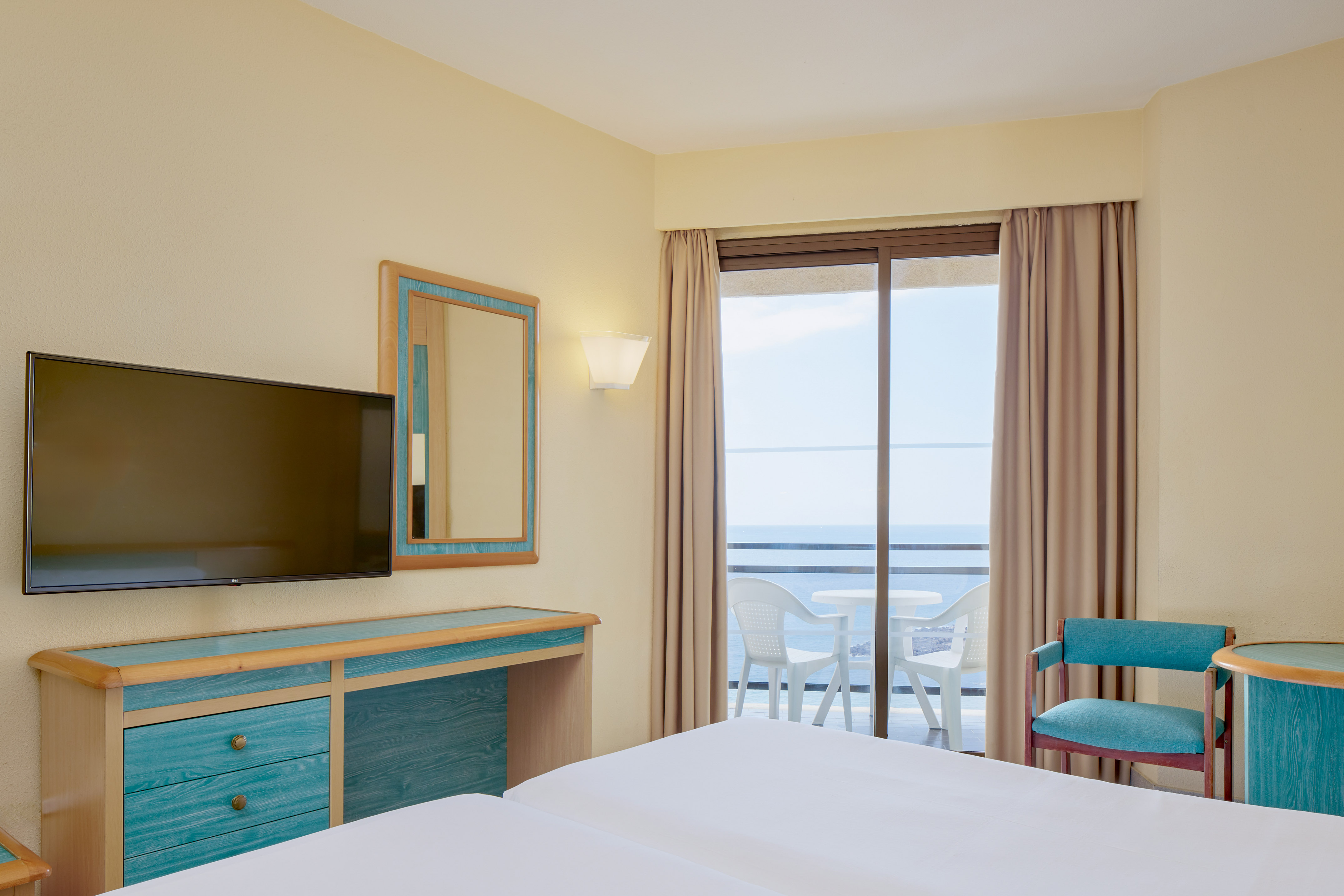 Hotel guest room with sea view balcony, flat-screen TV, and teal-accented desk.