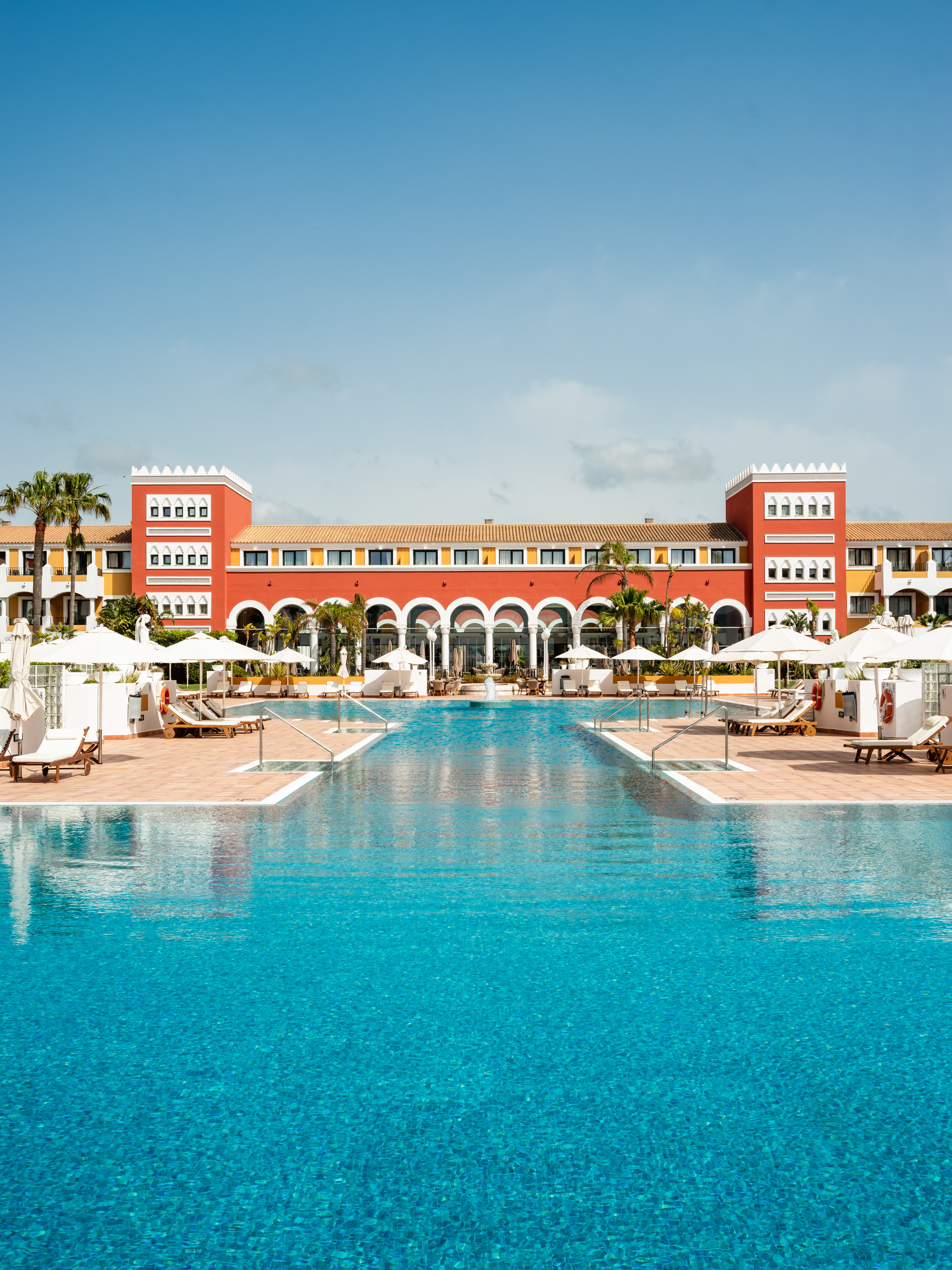 Red and yellow luxury hotel with large swimming pool and palm trees.