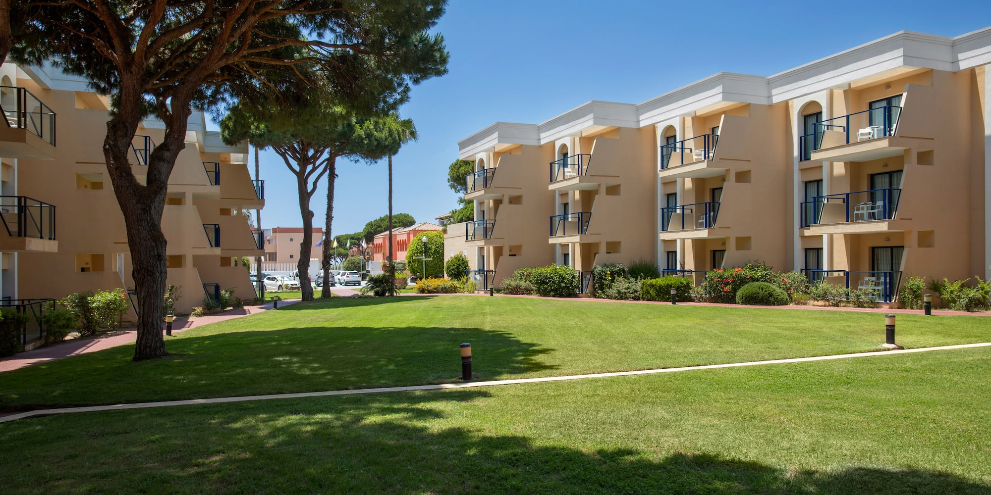 Sunny resort buildings with balconies, a lush green lawn, and tall trees.