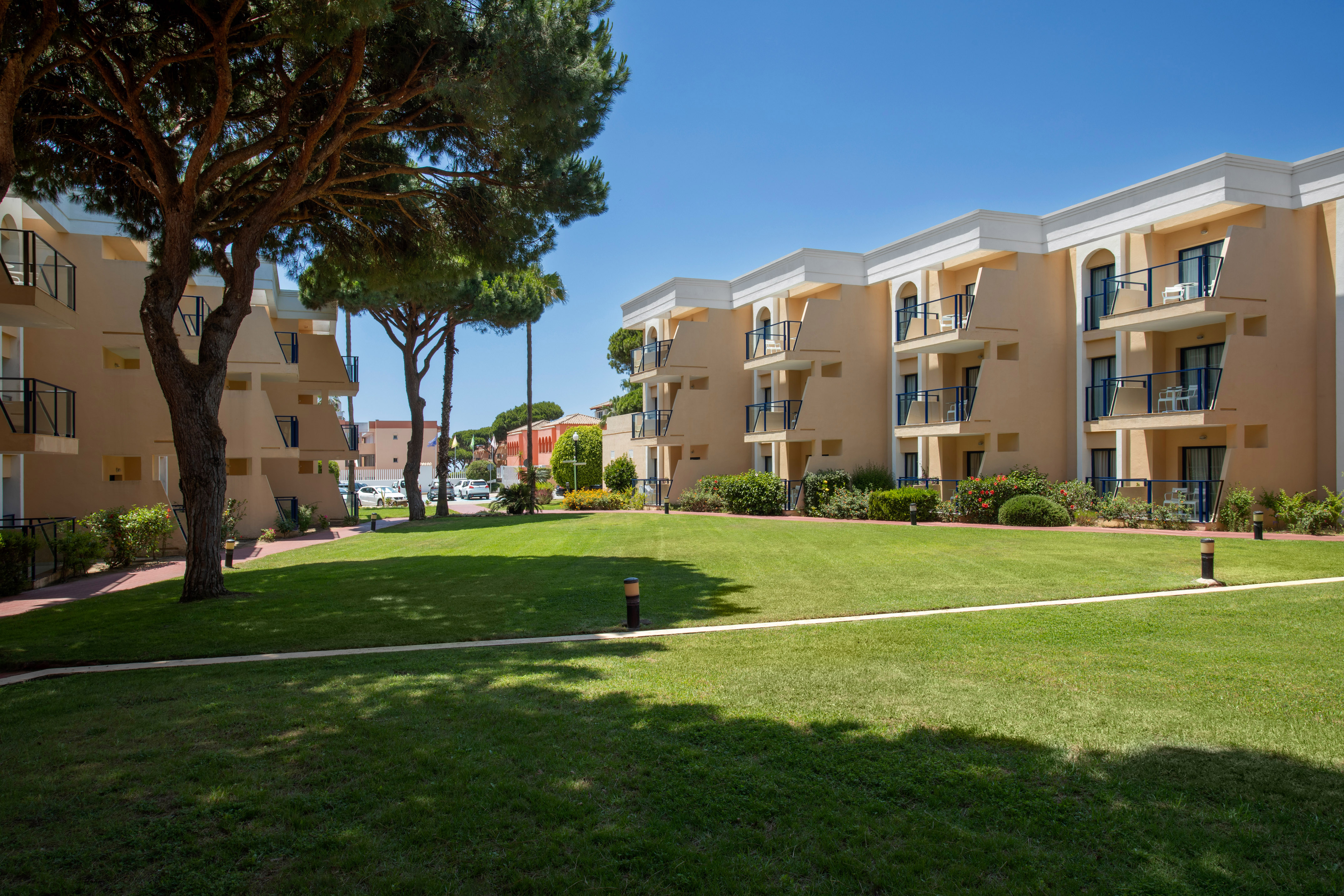 Sunny resort buildings with balconies, a lush green lawn, and tall trees.
