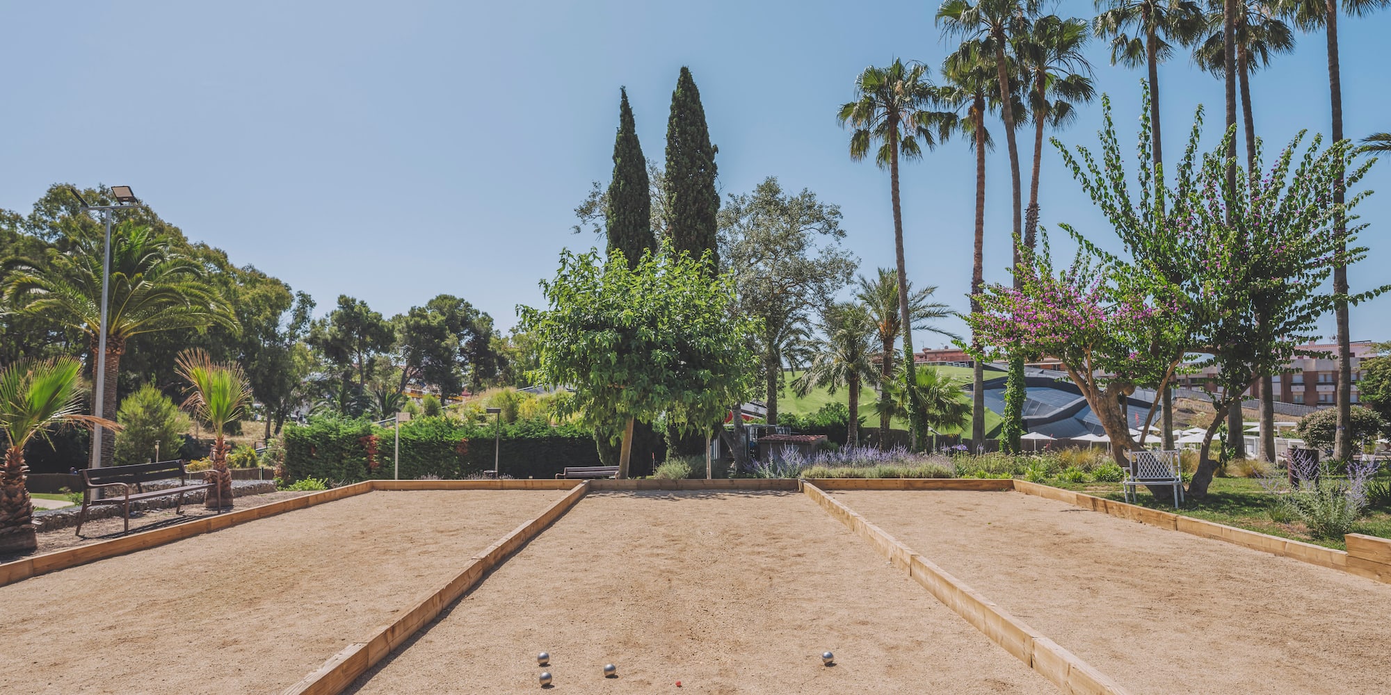 Sandy pétanque court with balls, surrounded by palm trees and greenery.