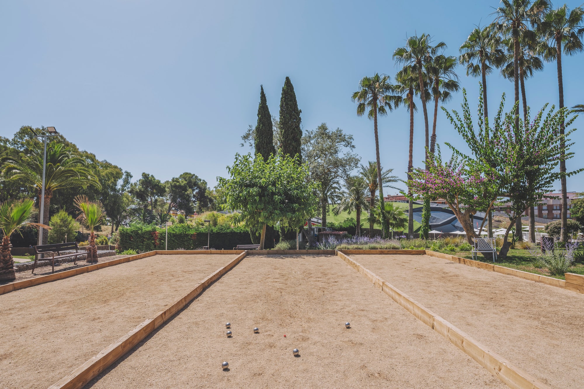 Sandy pétanque court with balls, surrounded by palm trees and greenery.
