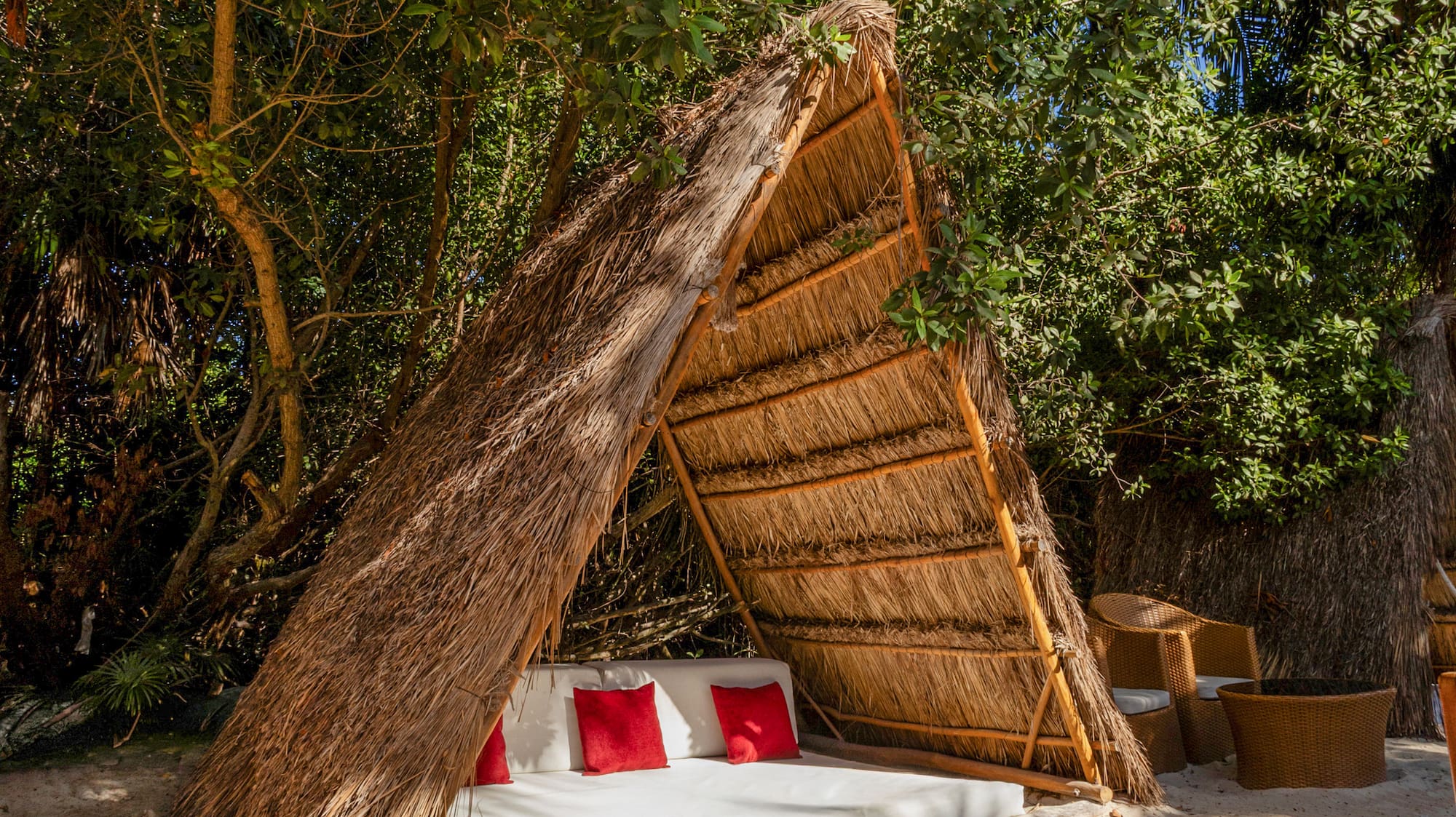 Relaxing beach cabana with straw roof, white bed, red pillows on sand.