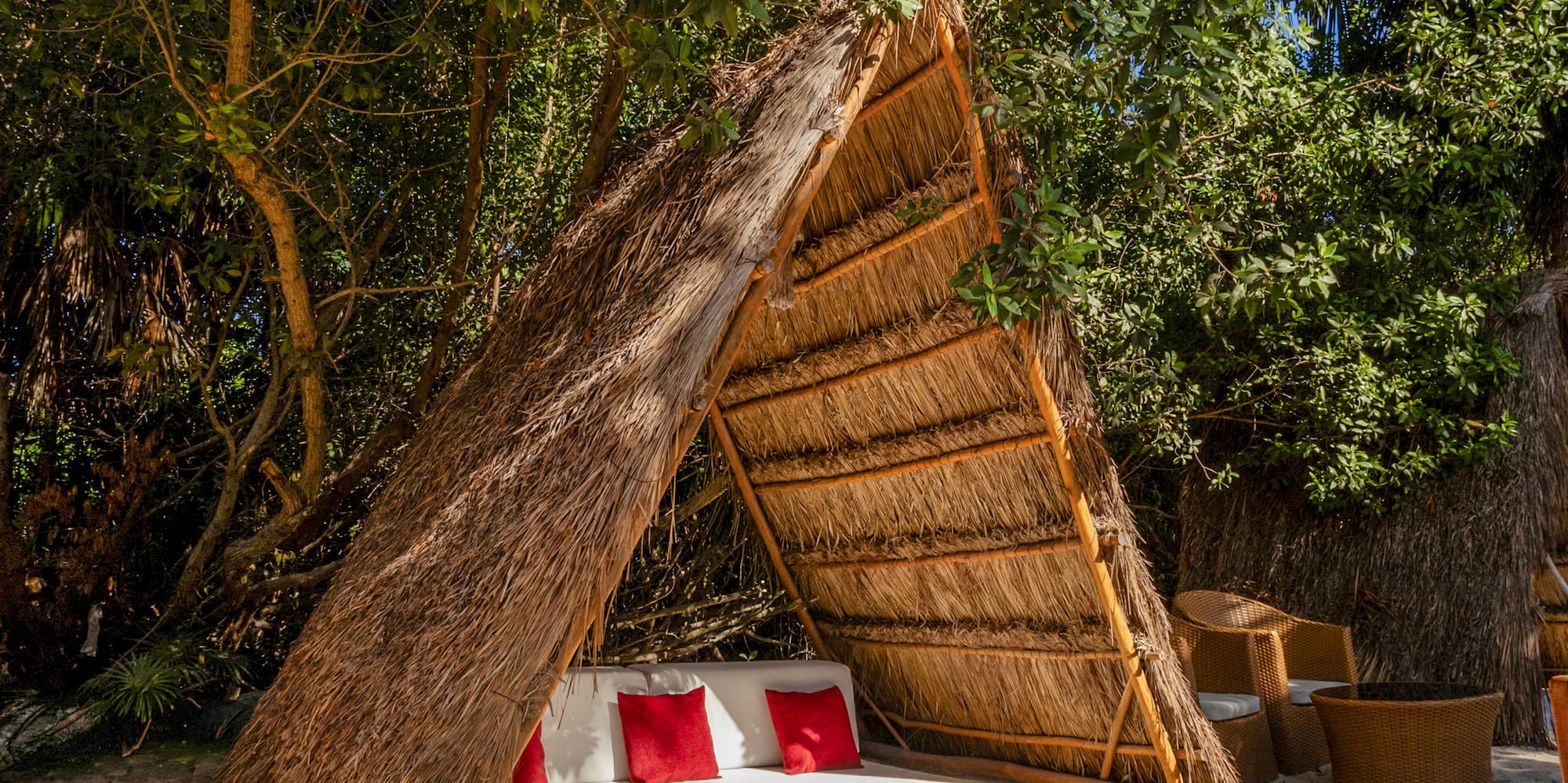 Relaxing beach cabana with straw roof, white bed, red pillows on sand.