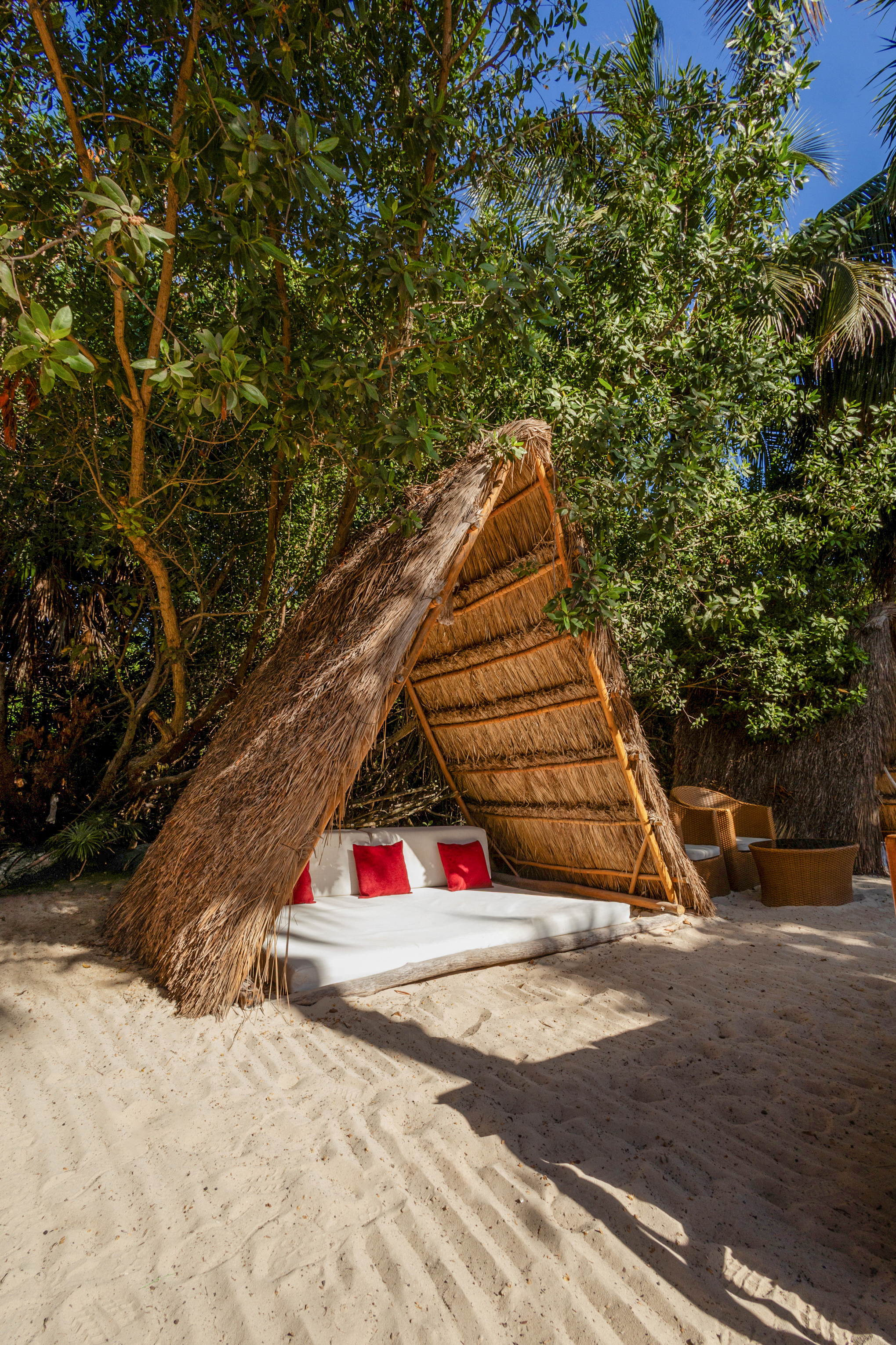 Relaxing beach cabana with straw roof, white bed, red pillows on sand.