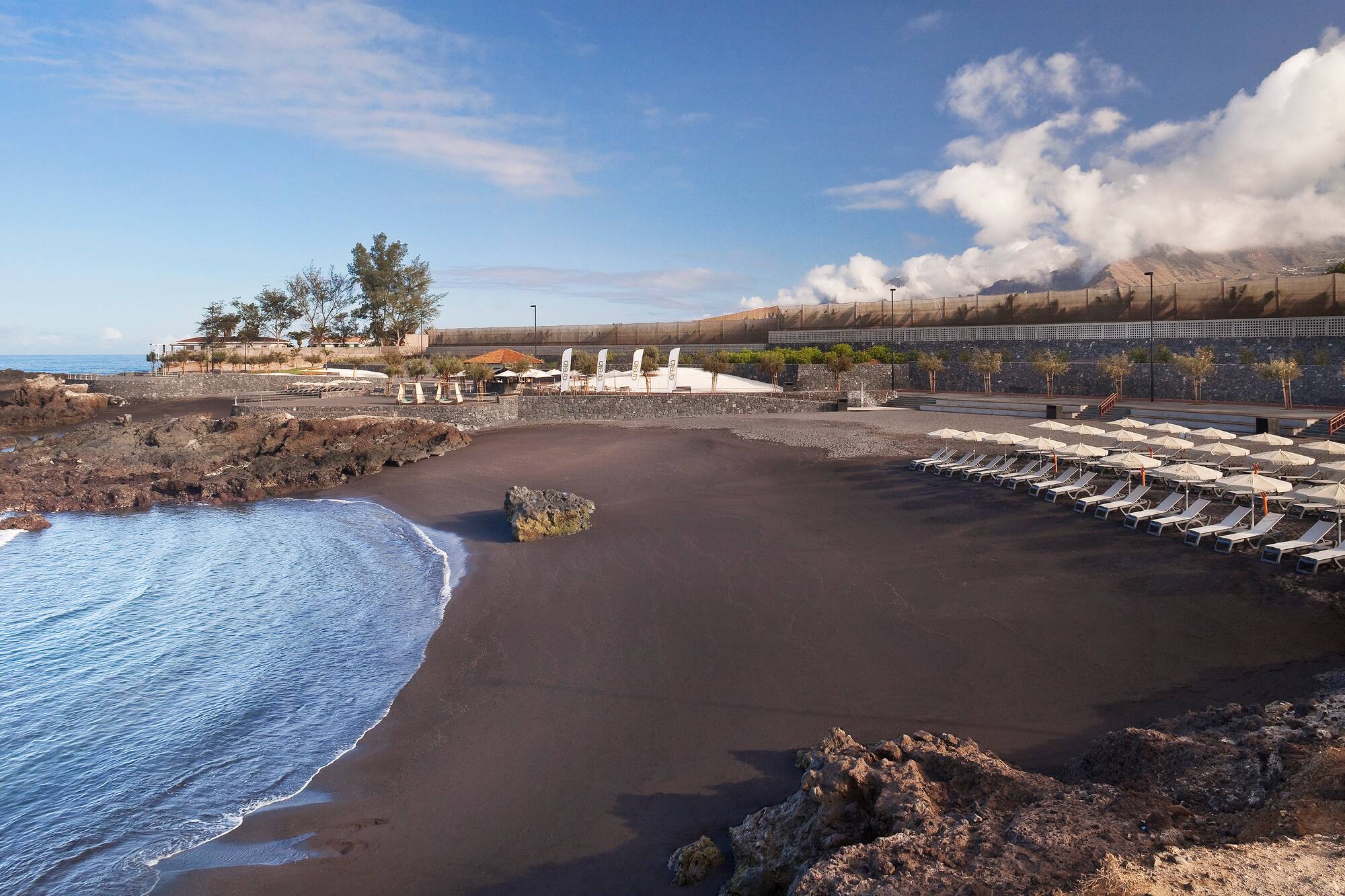 Volcanic black sand beach with ocean waves, sun loungers, and rocky landscape.