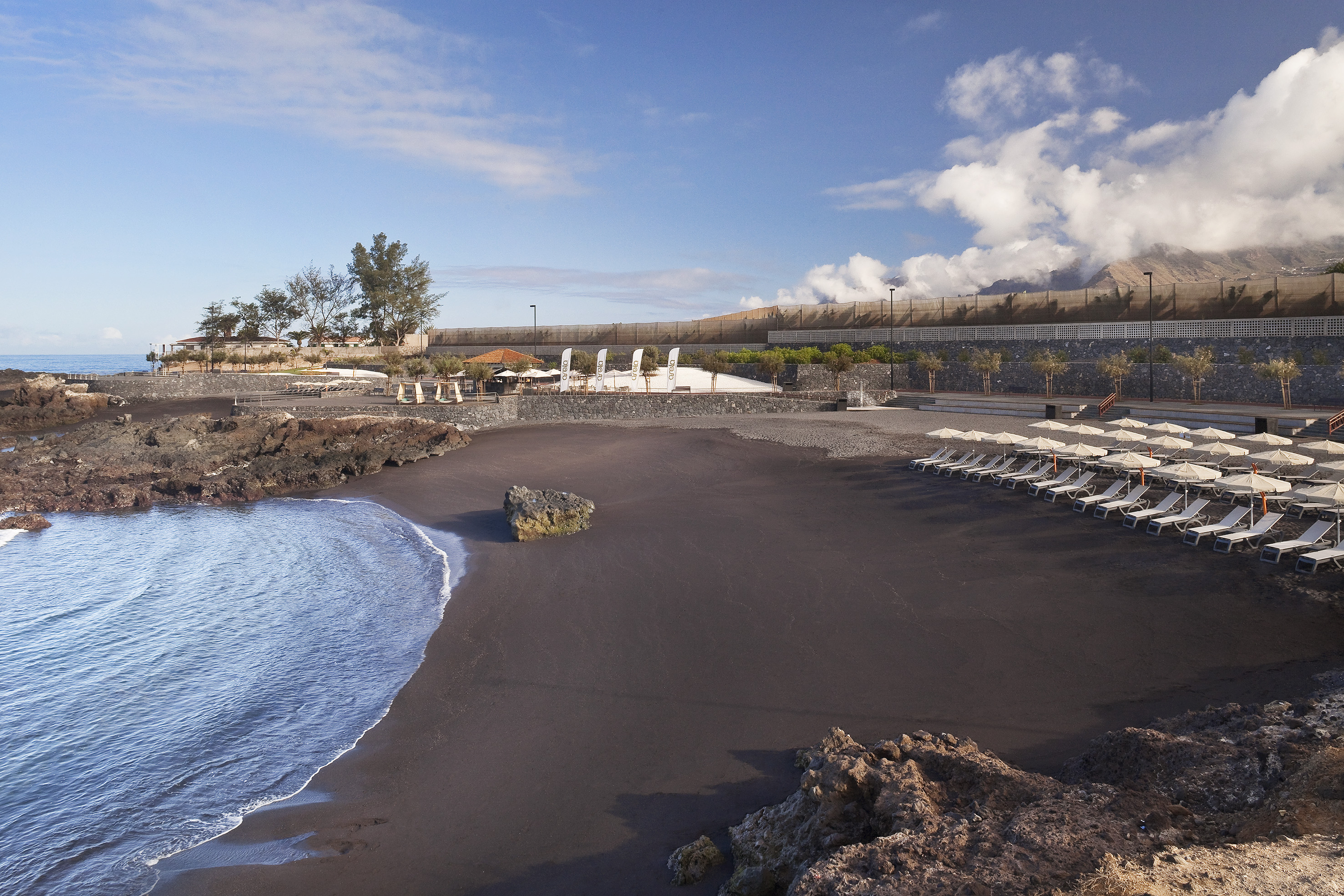 Volcanic black sand beach with ocean waves, sun loungers, and rocky landscape.