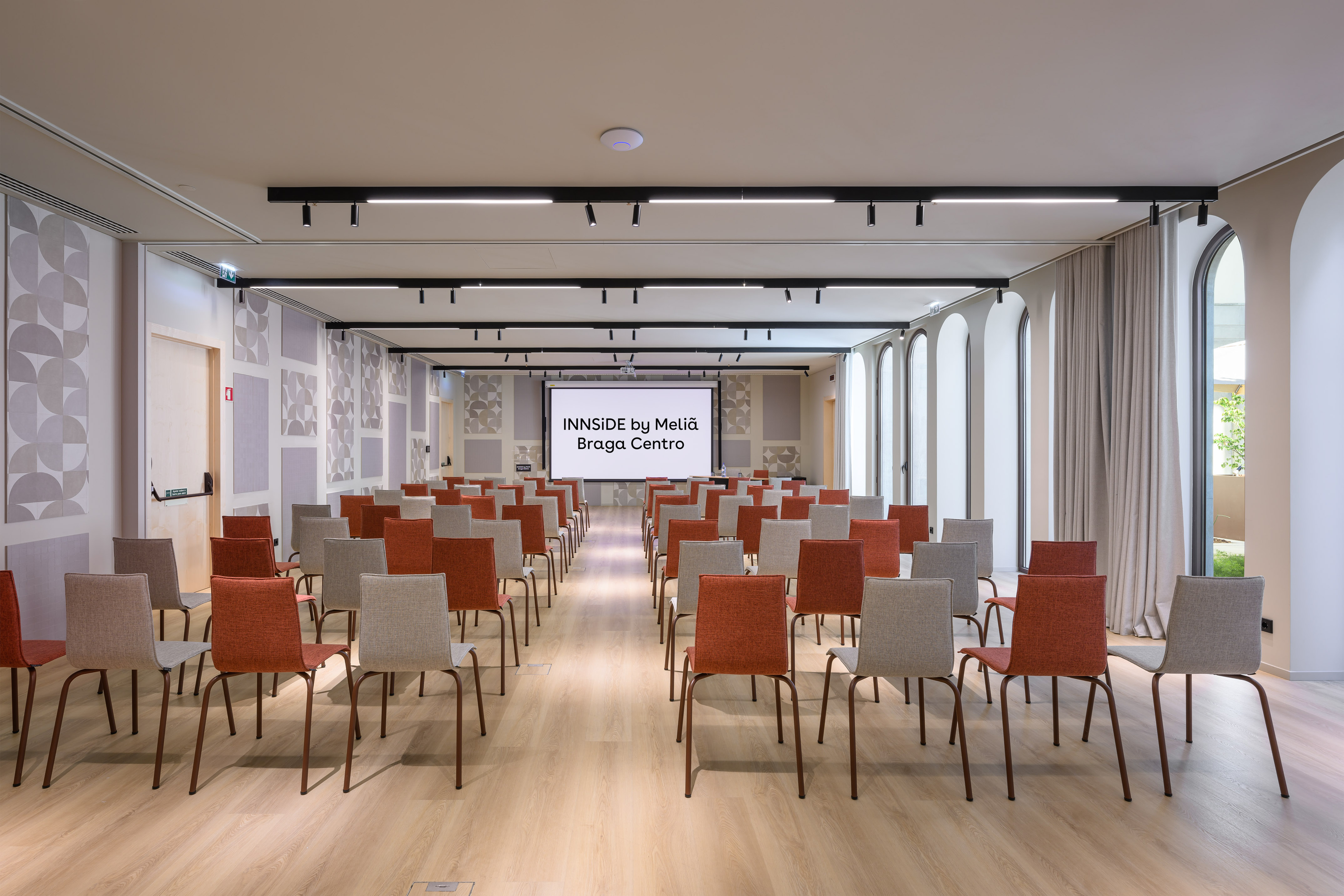 Modern conference room setup with red and grey chairs facing a presentation screen. - INNSiDE Braga Centro, Braga
