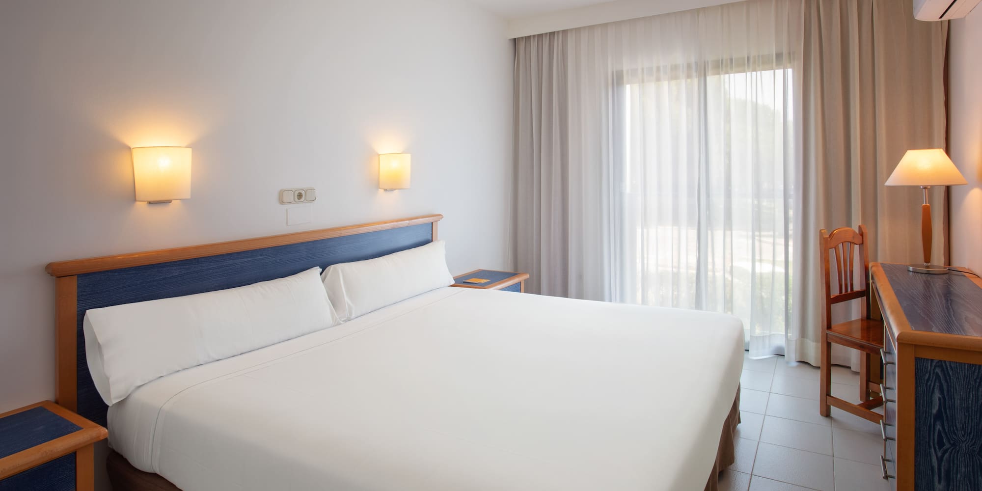 Bright hotel room with white bed, blue headboard, and large window.