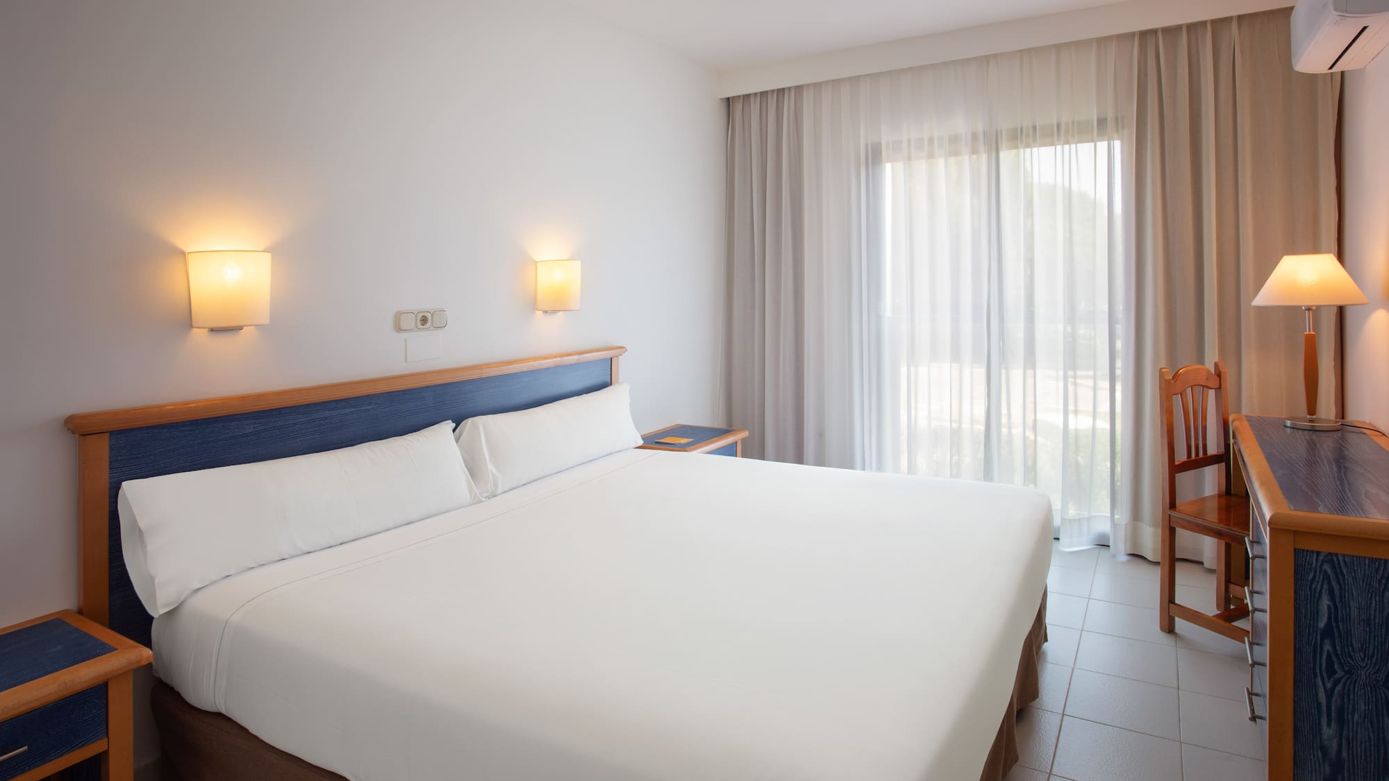 Bright hotel room with white bed, blue headboard, and large window.