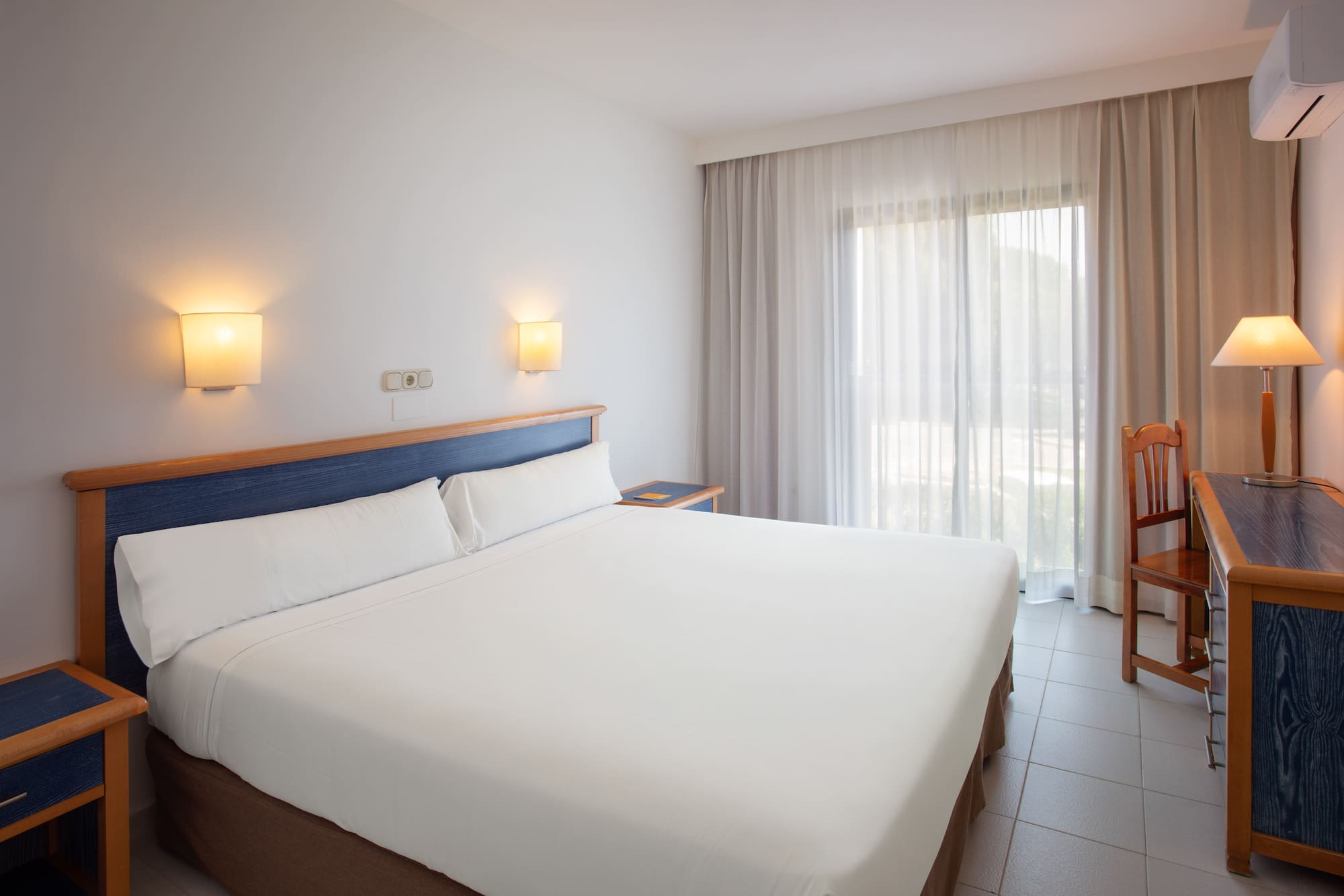 Bright hotel room with white bed, blue headboard, and large window.