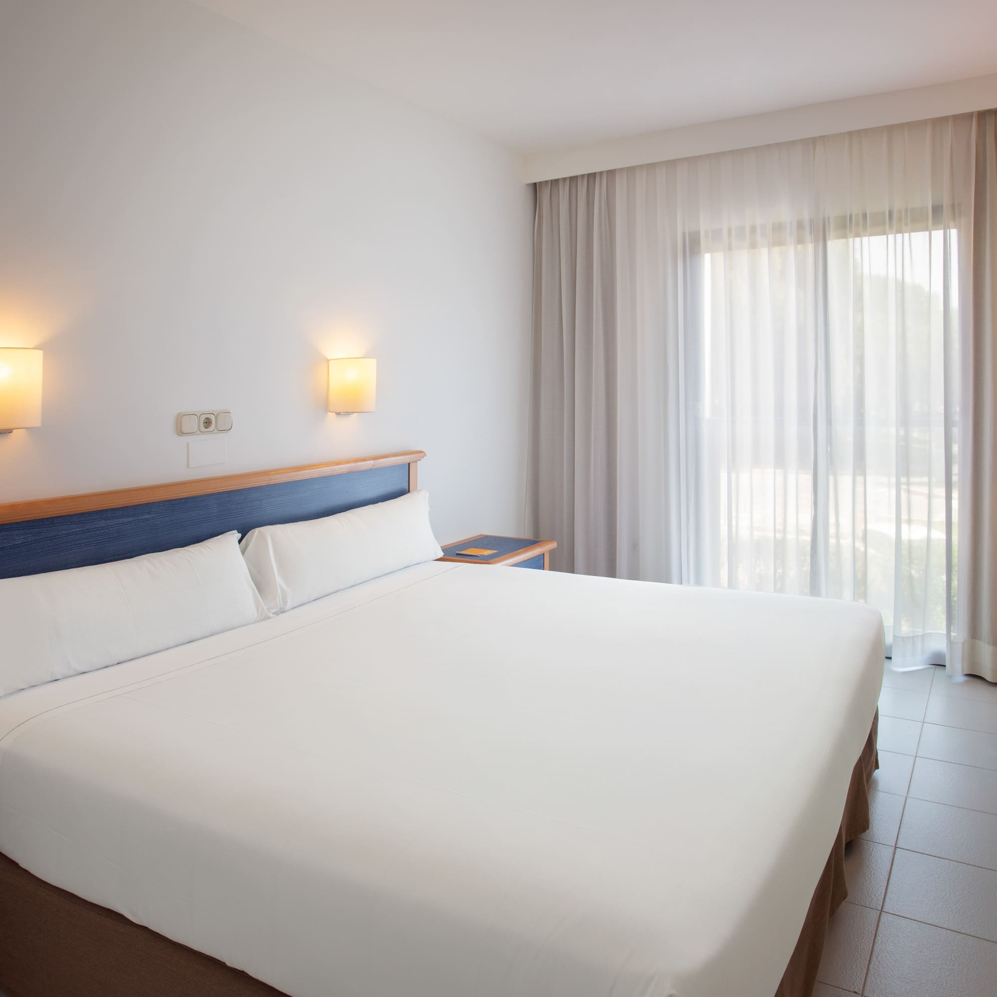Bright hotel room with white bed, blue headboard, and large window.