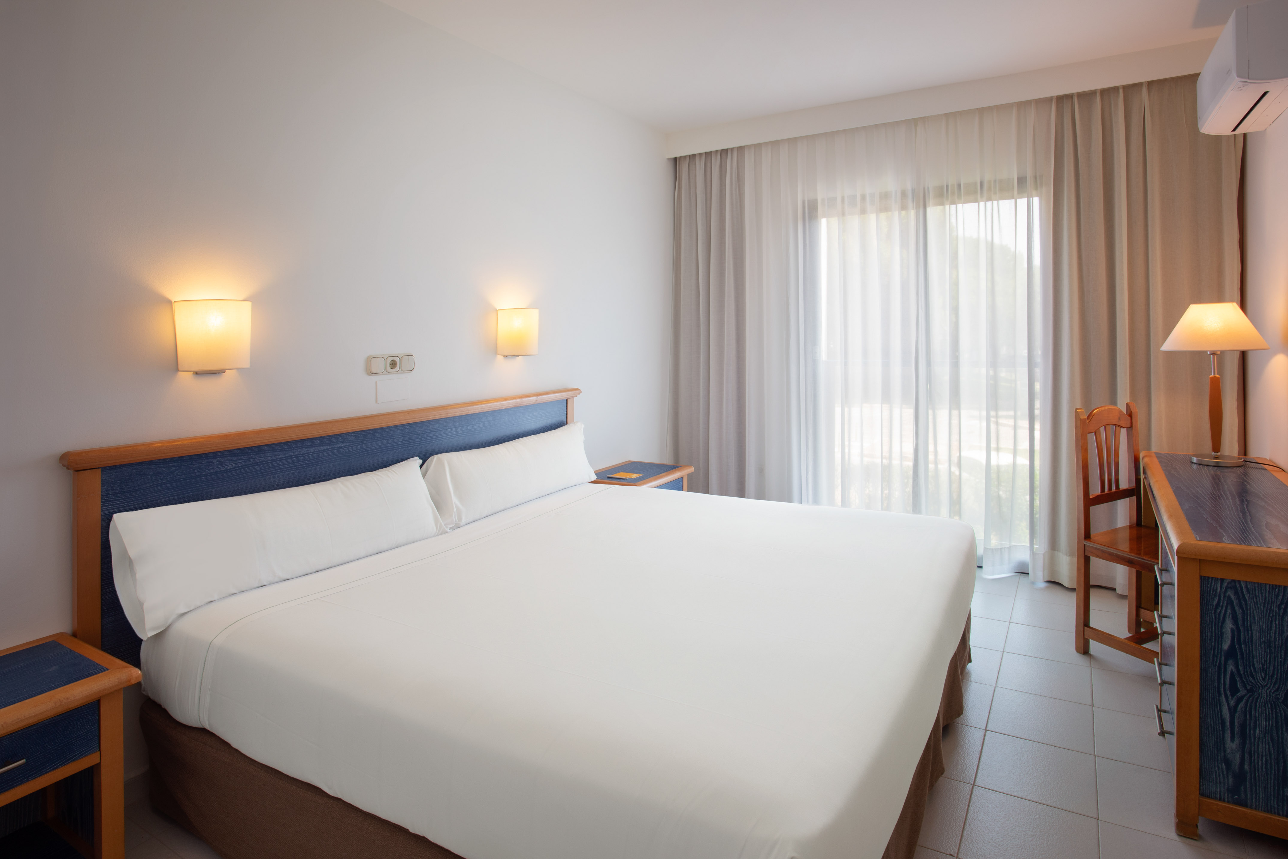 Bright hotel room with white bed, blue headboard, and large window.
