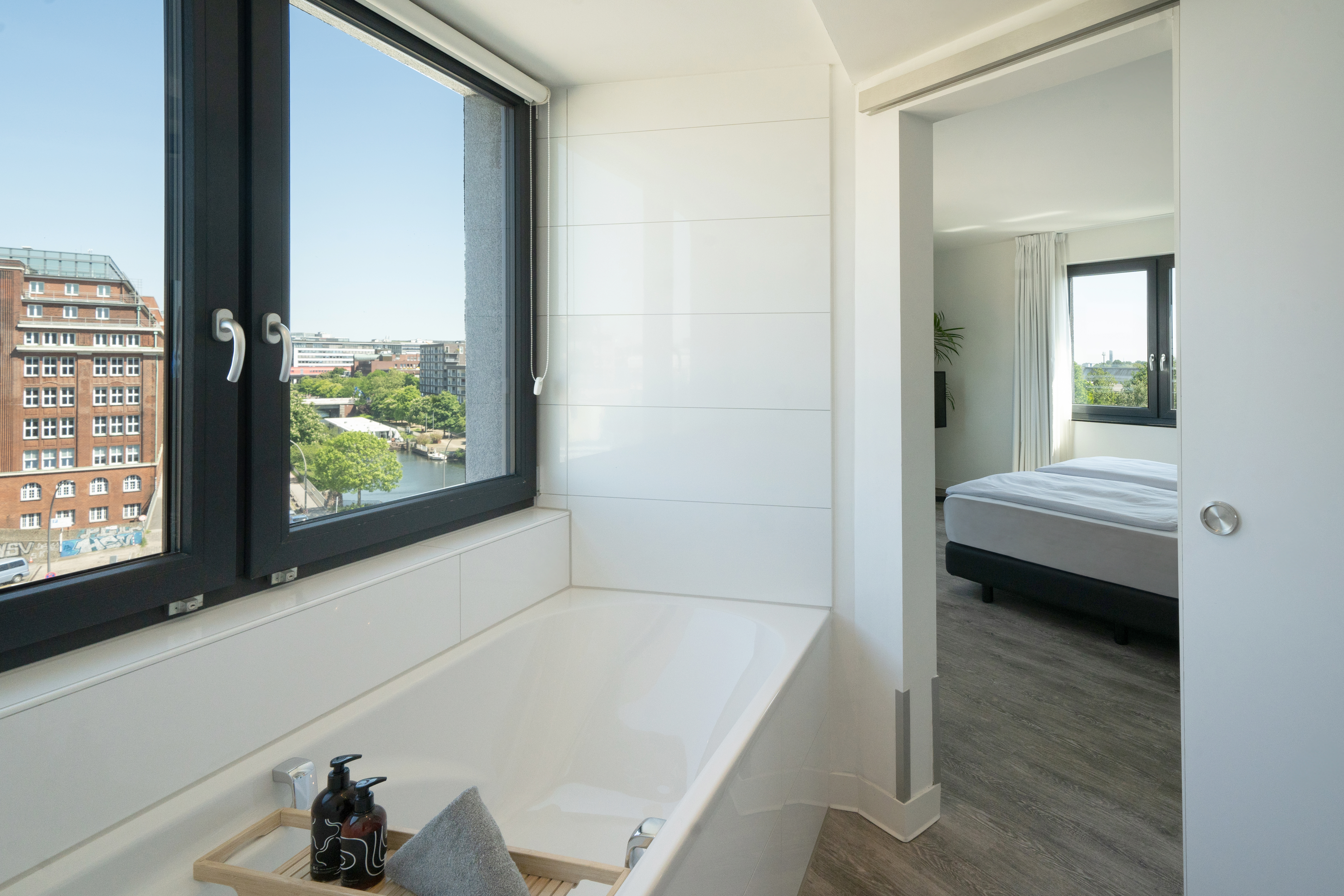 Modern bathroom with bathtub and city view, leading to bright bedroom.