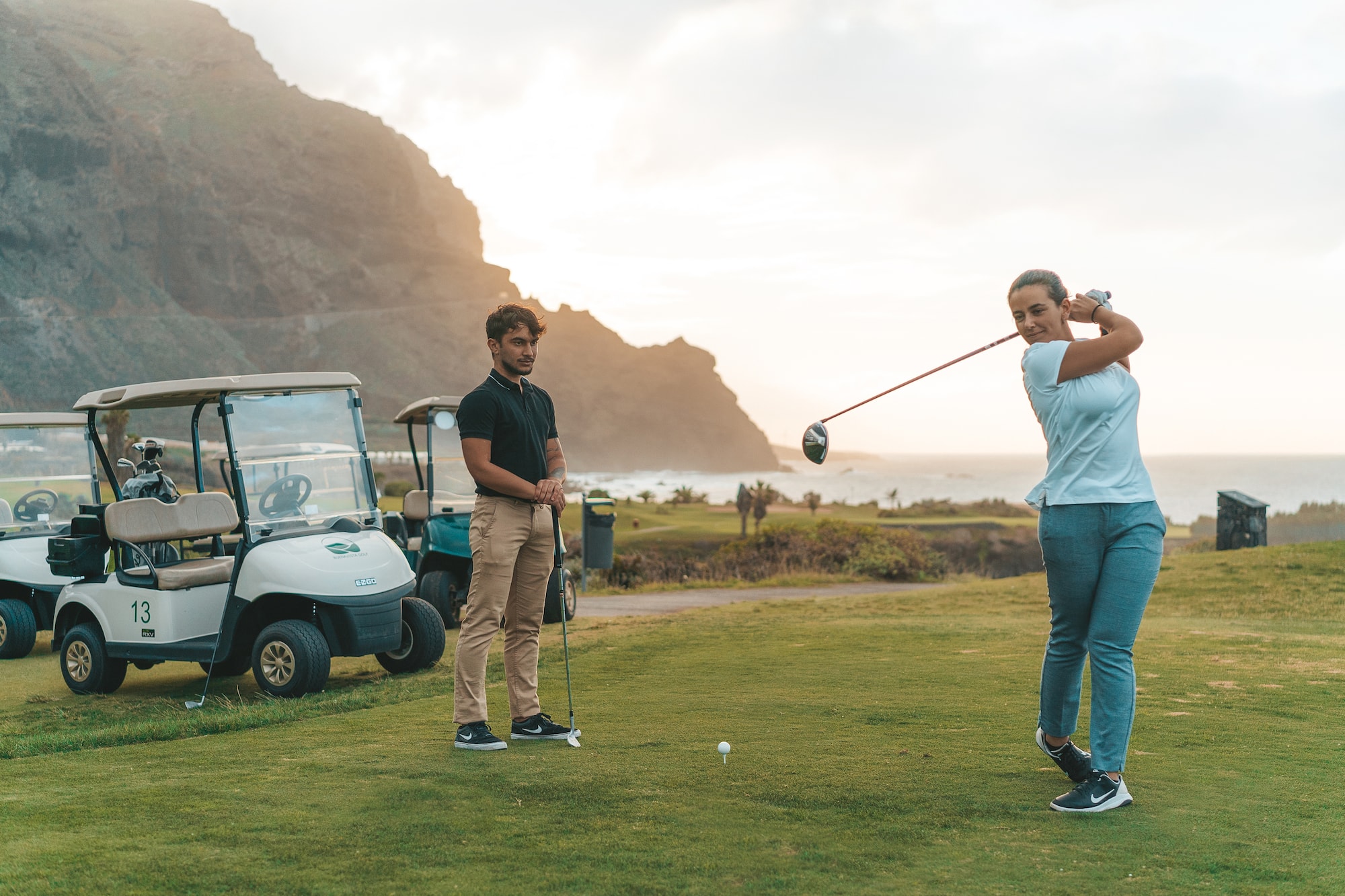 Female golfer swings driver on ocean view golf course, man and golf cart.