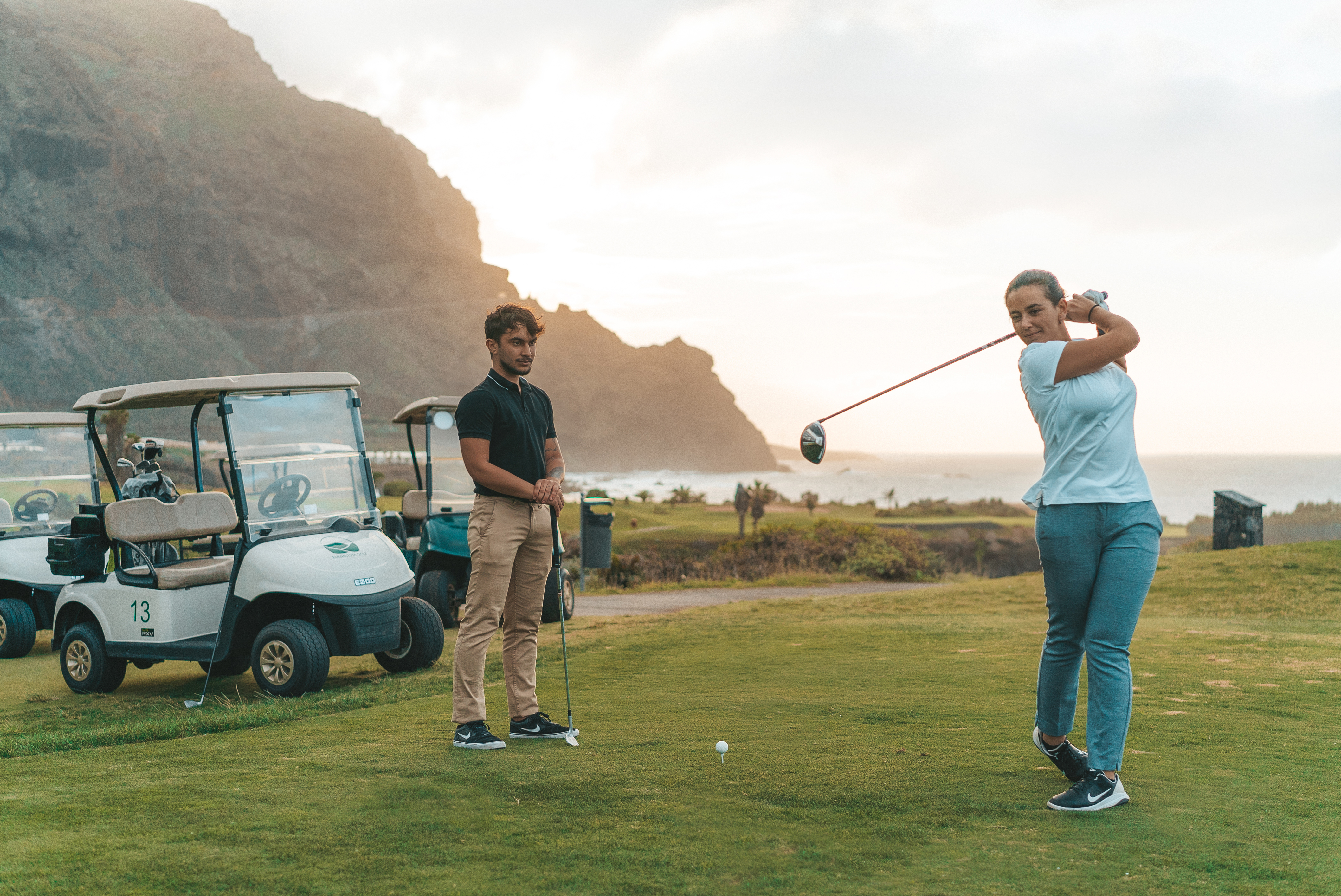 Female golfer swings driver on ocean view golf course, man and golf cart.
