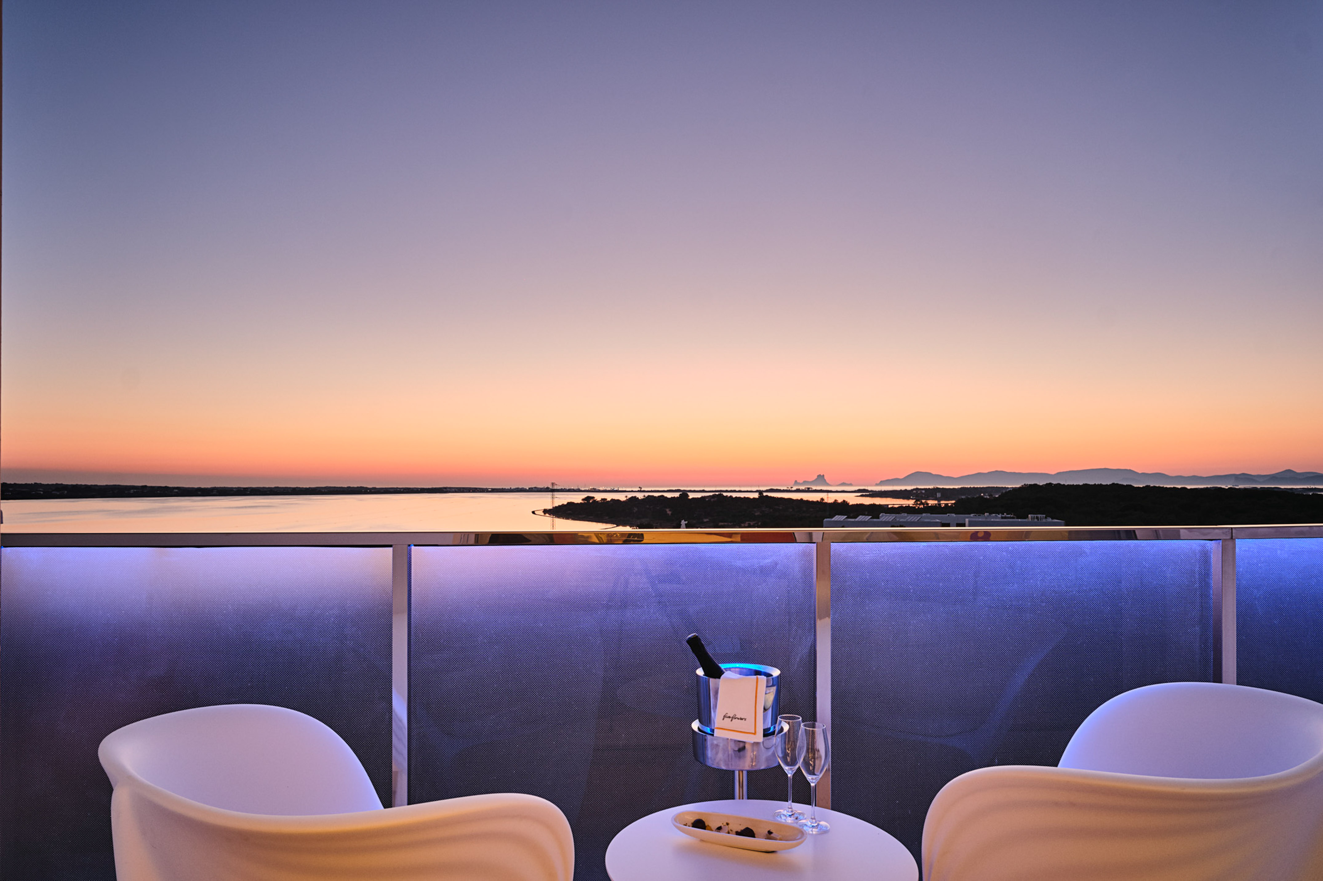 Luxury balcony sunset view with champagne, ocean, and lighted railing.