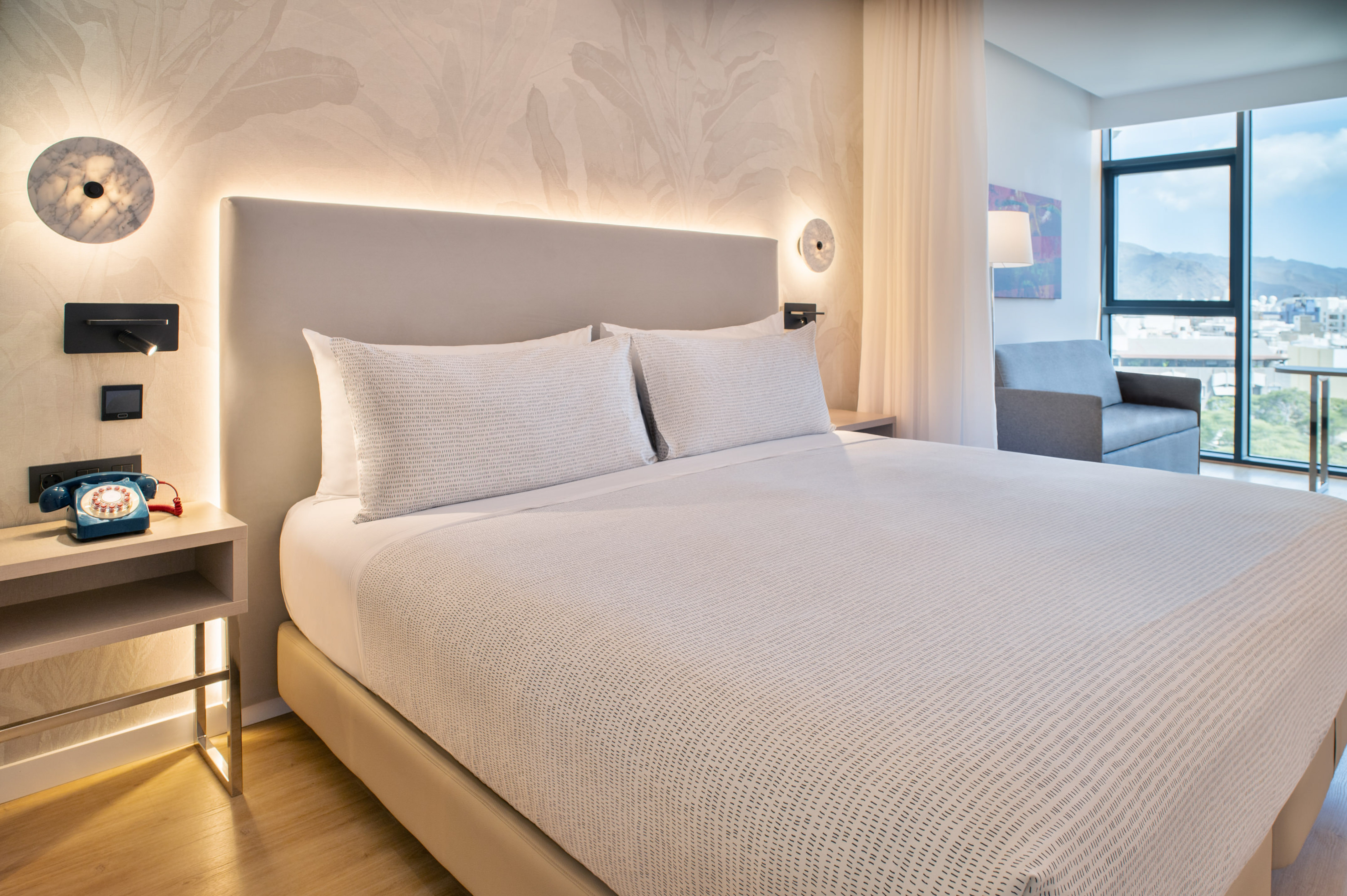 Modern hotel bedroom with king bed, illuminated headboard, city window view.