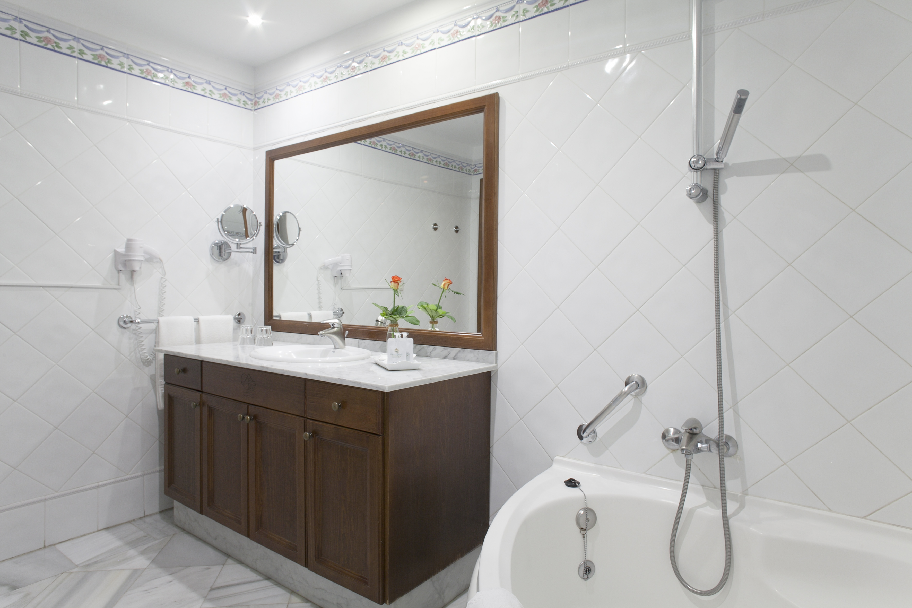 Hotel bathroom with white tiles, wood vanity, sink, mirror, and bathtub-shower.