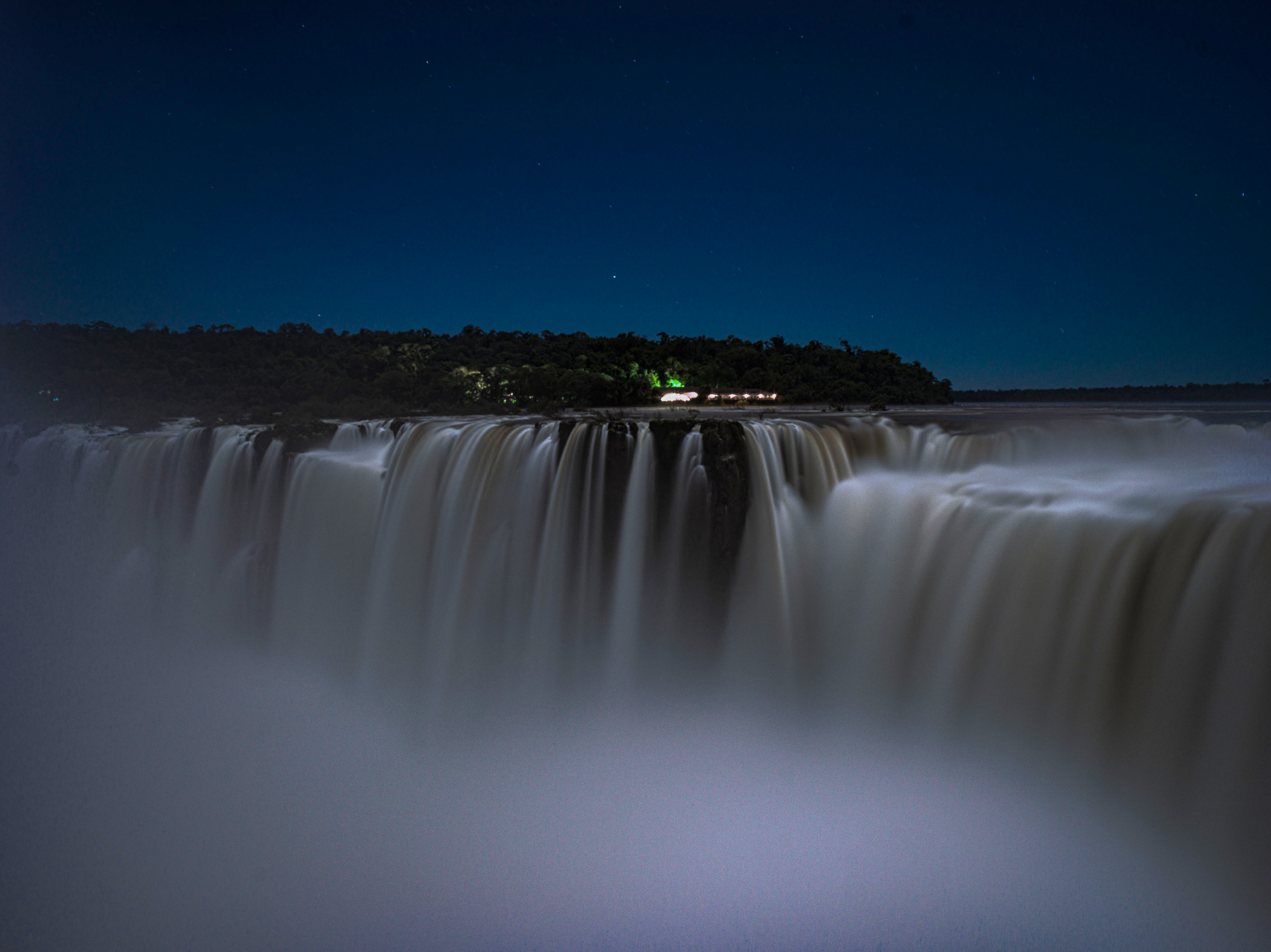 Iguazu Falls at night under starry sky, jungle, and misty water.