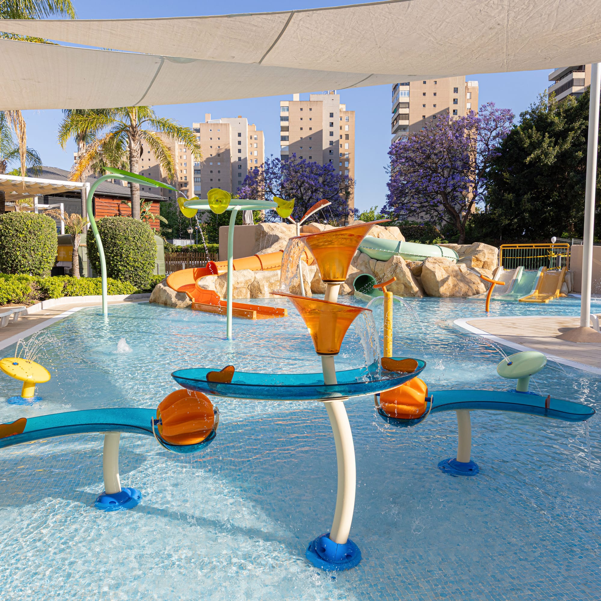 Sunny resort water park with slides, splash pad, palm trees, and hotel buildings.