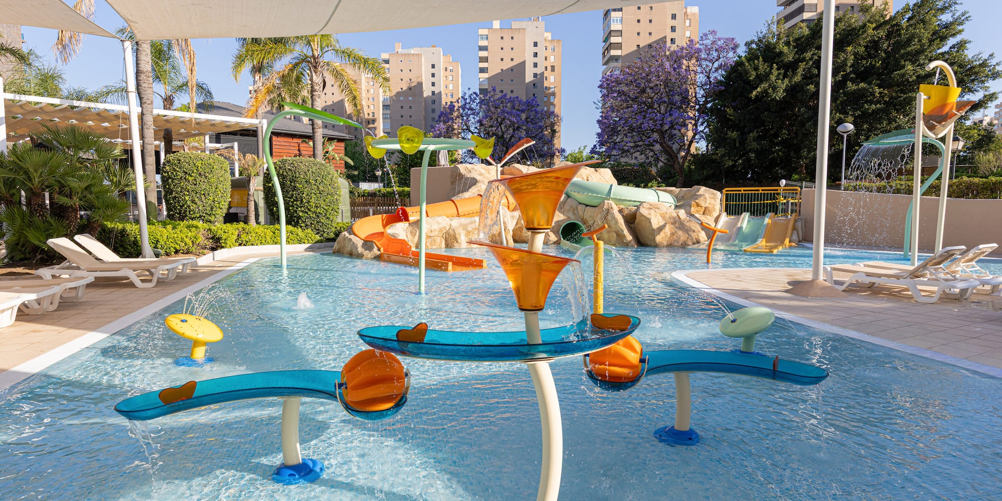 Sunny resort water park with slides, splash pad, palm trees, and hotel buildings.