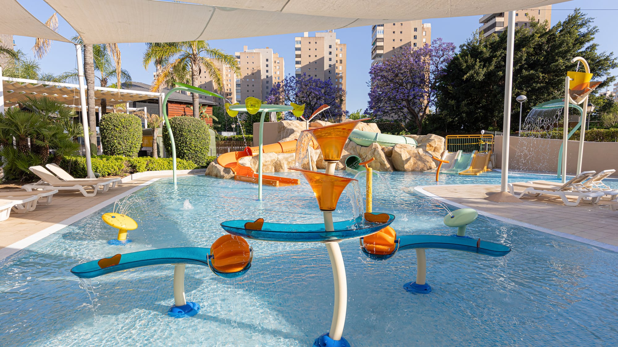 Sunny resort water park with slides, splash pad, palm trees, and hotel buildings.