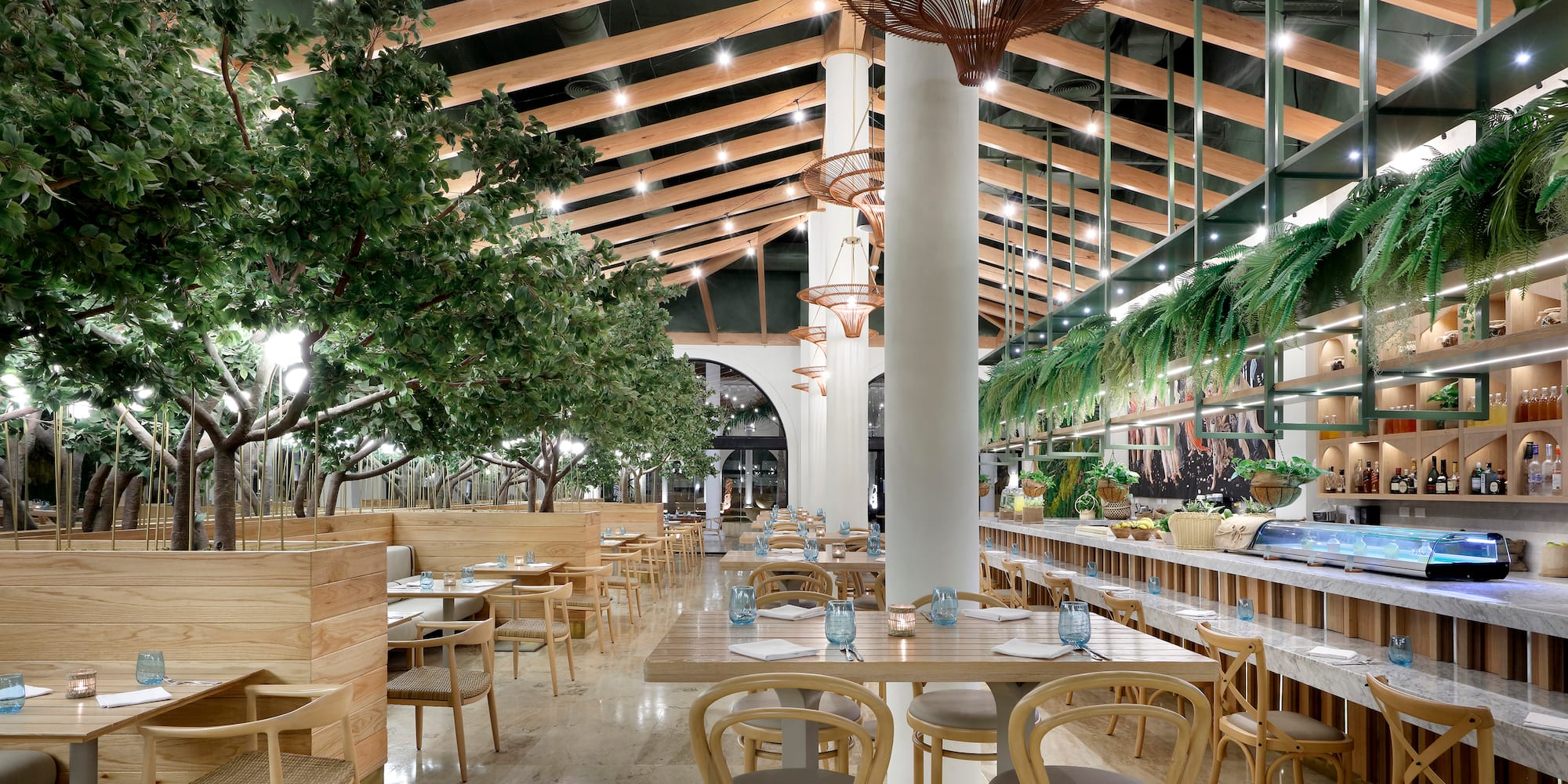 Modern restaurant with indoor trees, wood ceilings, dining tables, and bar.