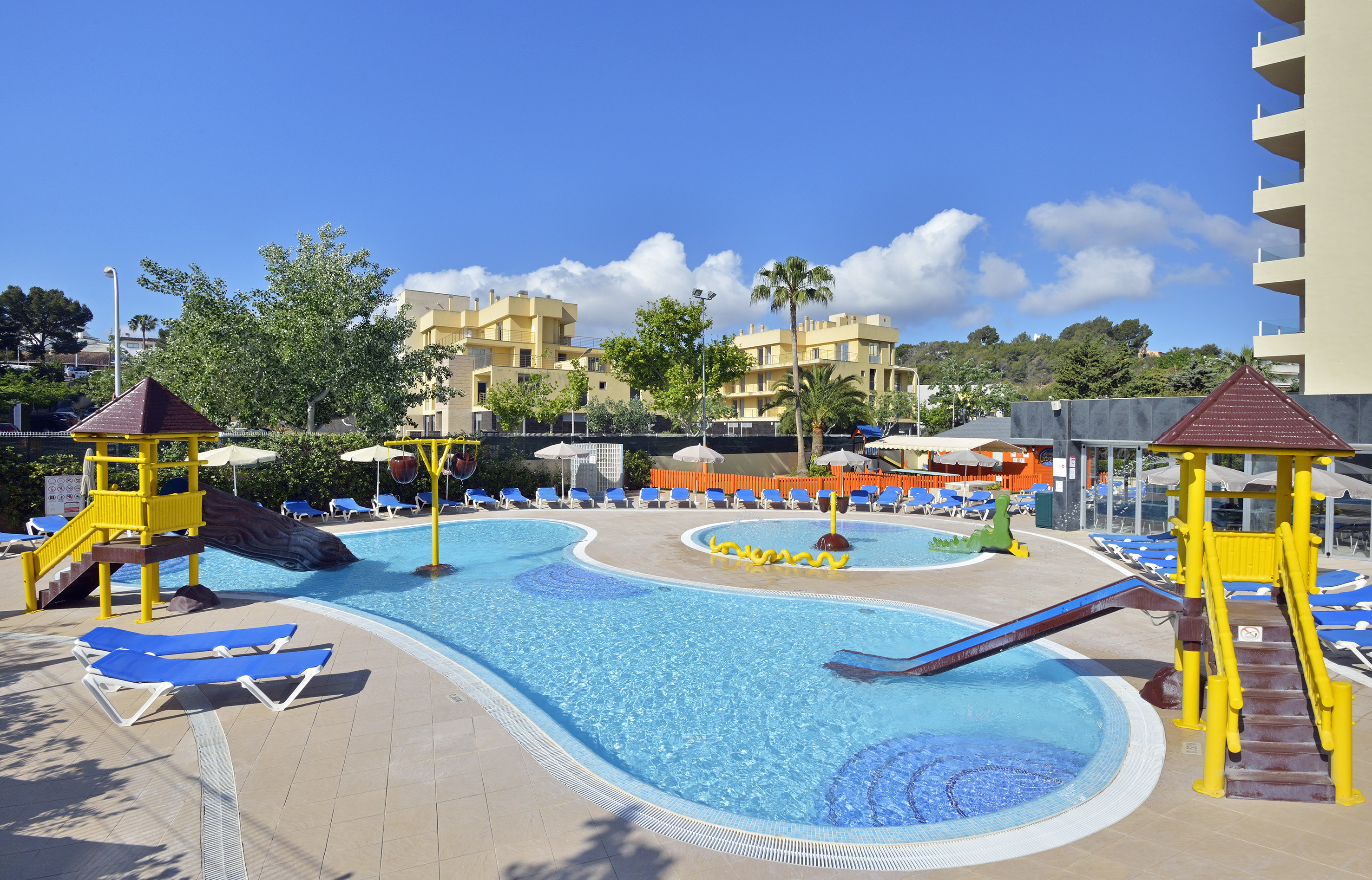 Sunny resort family pool with water slides, play areas, and lounge chairs.