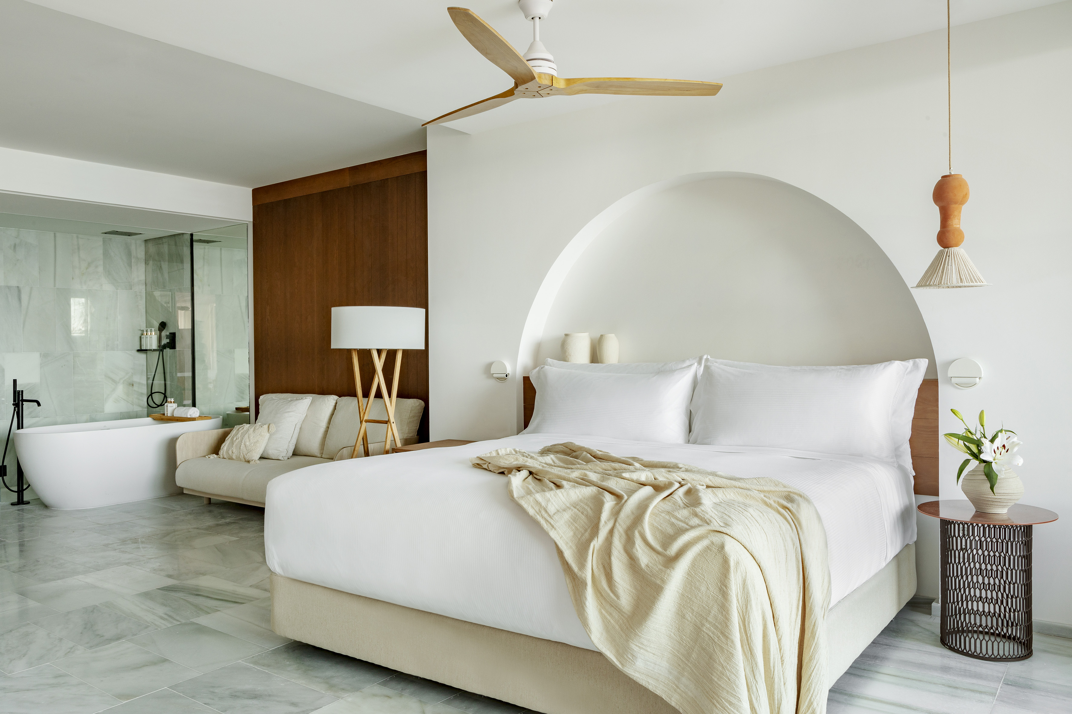 Modern hotel bedroom with white bed, arch, marble floor, and open luxury bath.