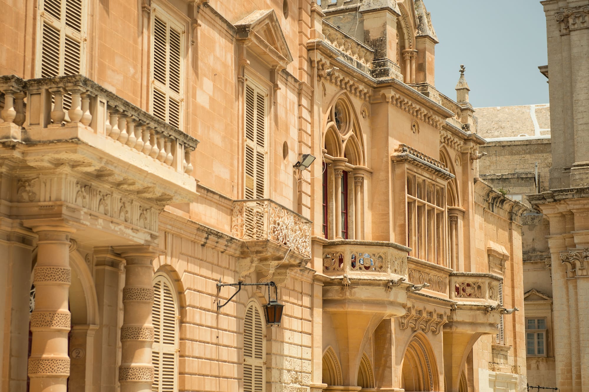 Ornate historic Maltese stone building with balconies and shuttered windows.